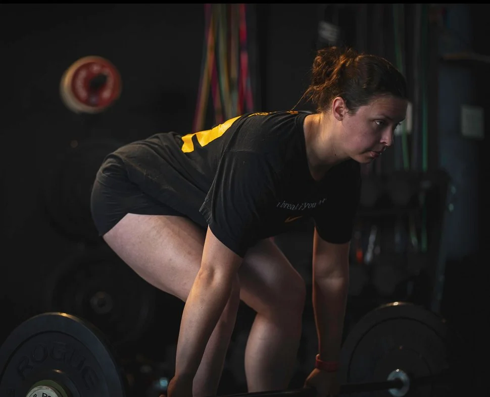 Young woman with dark hair tied in a bun, wearing a black t-shirt with yellow details and black shorts, lifting a barbell in a gym.