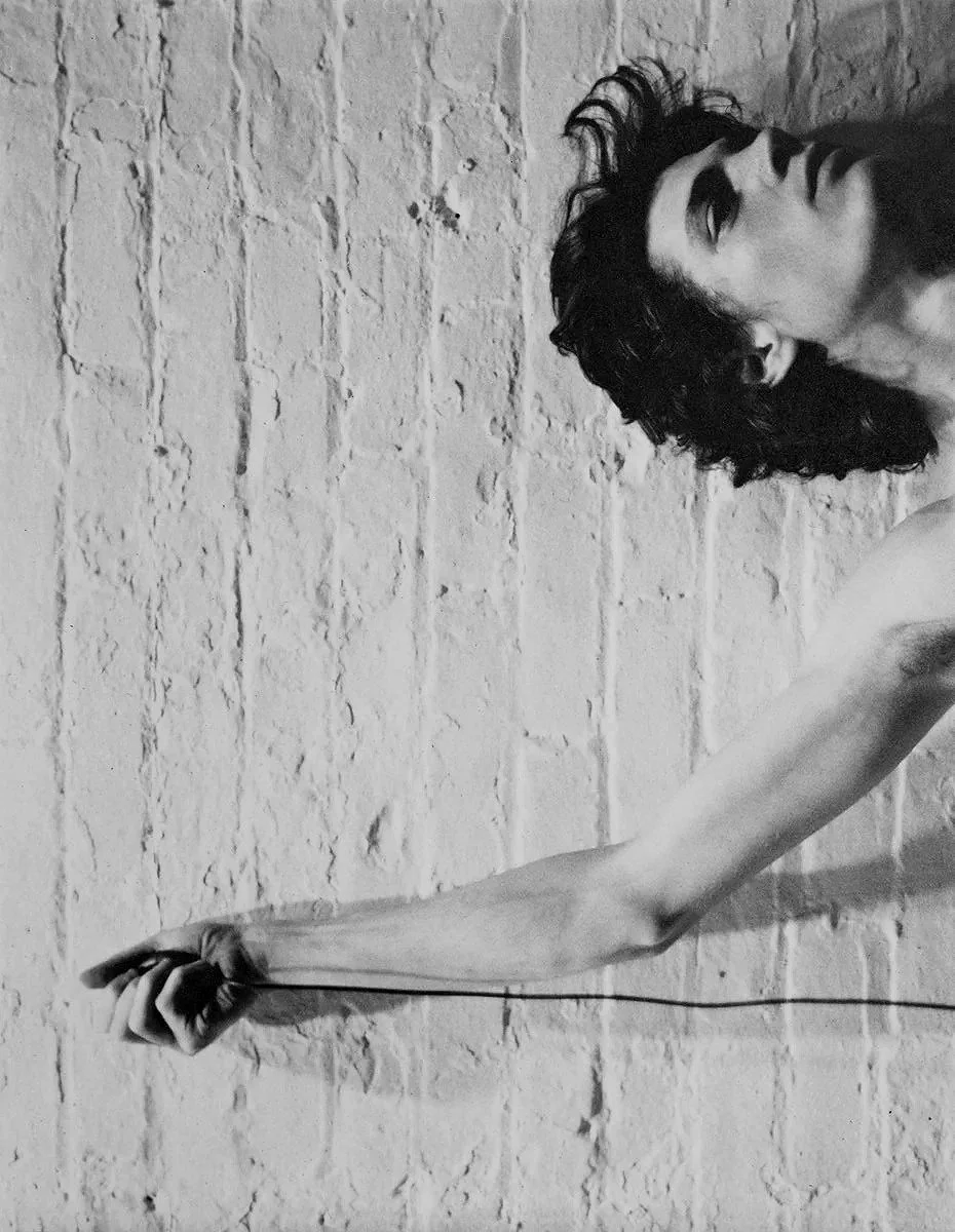 A black and white photo of a shirtless young man with long hair, wearing dark jeans, a chain necklace, and a bracelet, against a plain background.