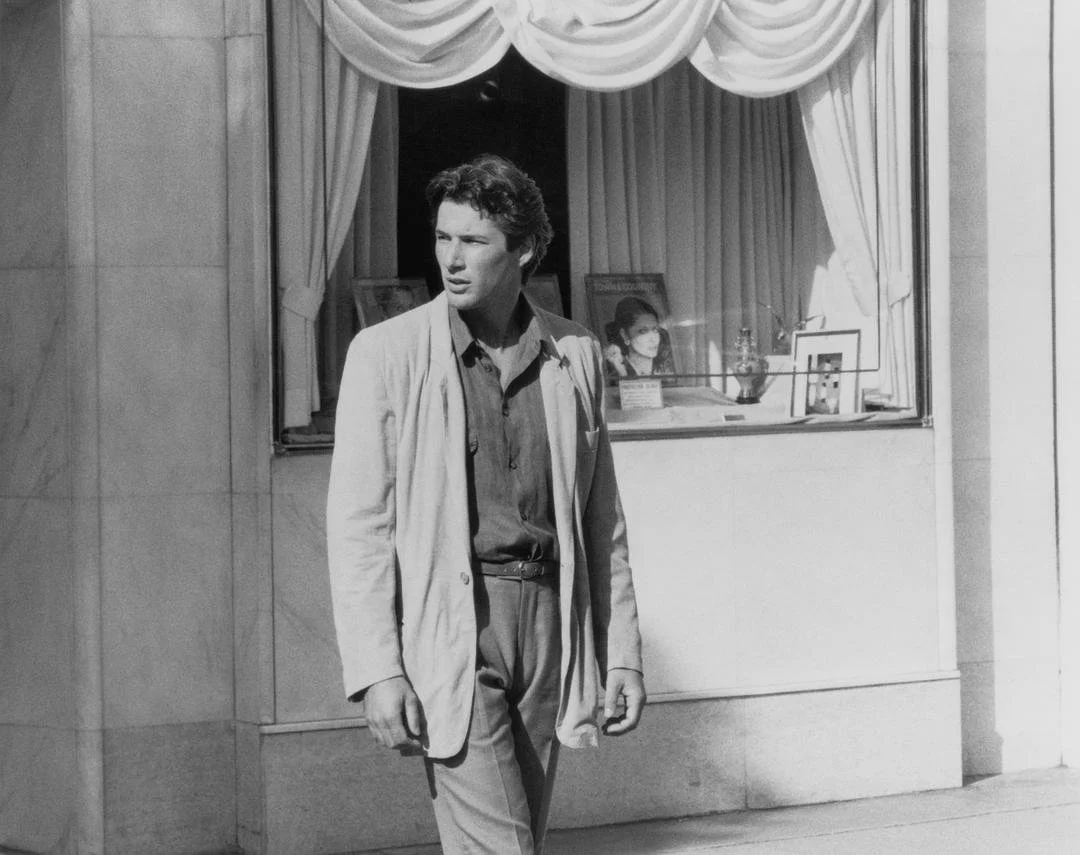 A black and white photograph of a man in profile, sitting with a serious expression, wearing a sleeveless, sparkly top, against a plain background.