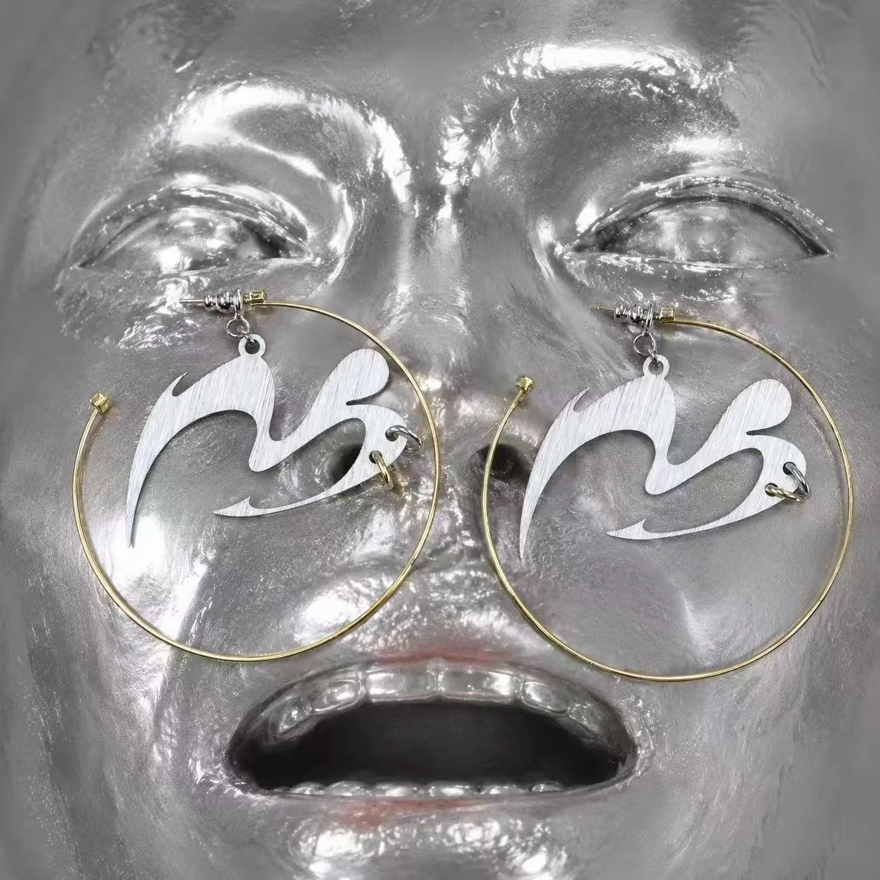 Silver and gold hoop earrings with a stylized abstract design, displayed on a reflective face-shaped background.