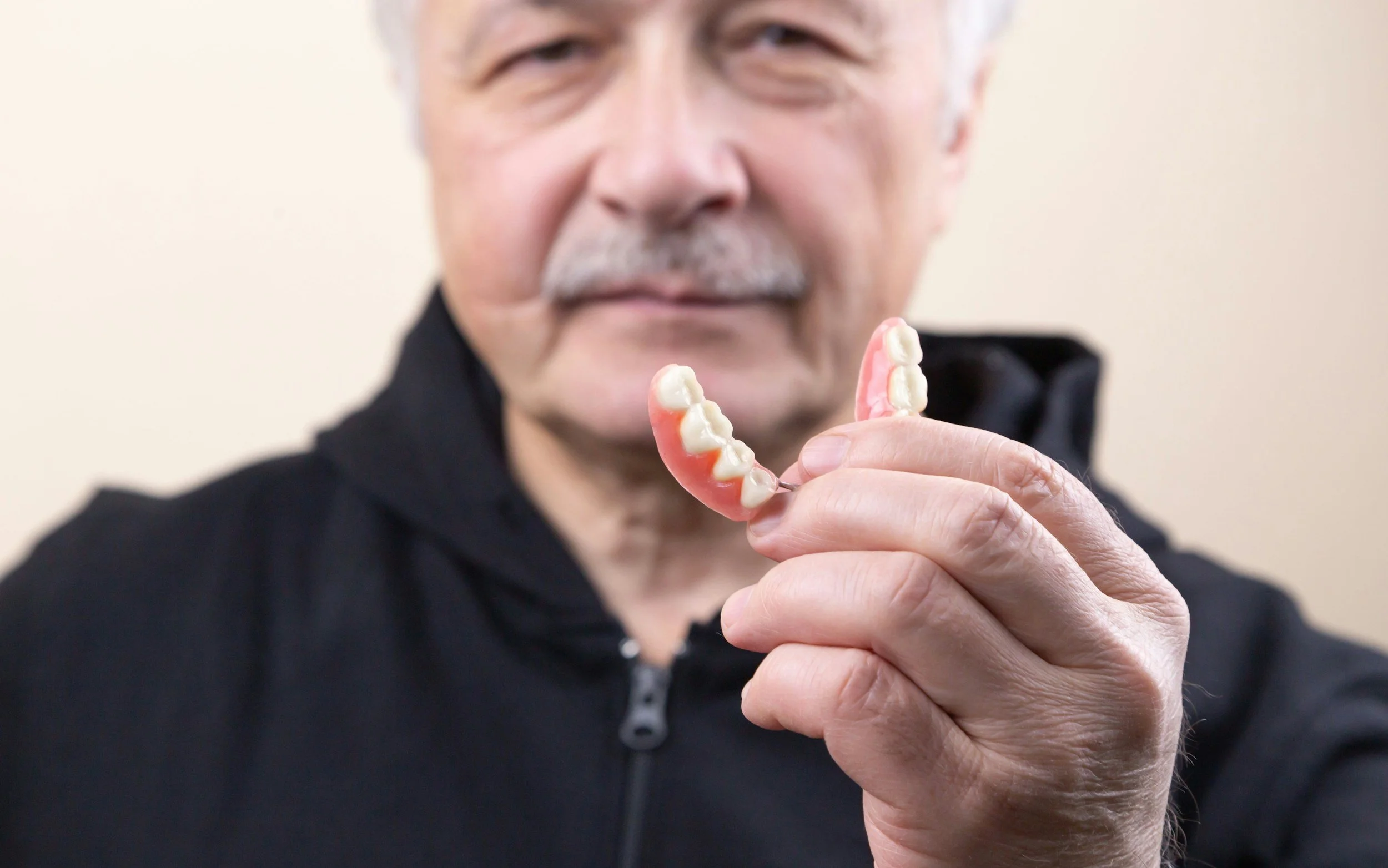 An older man holding a pink and white partial dentures towards the camera, with a blurred background.