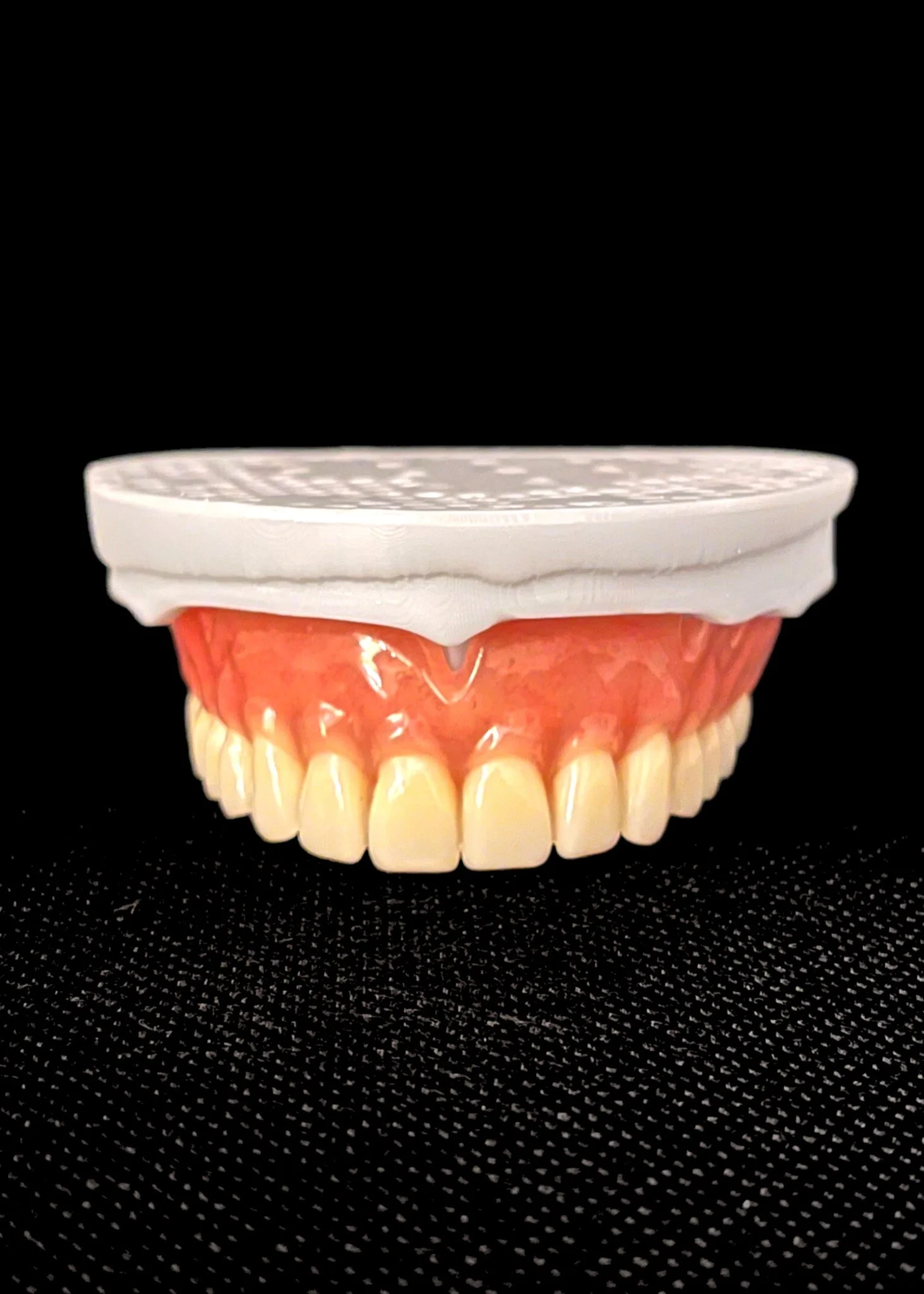 A set of artificial teeth and gums with a denture box lid on top, placed on a black fabric surface.