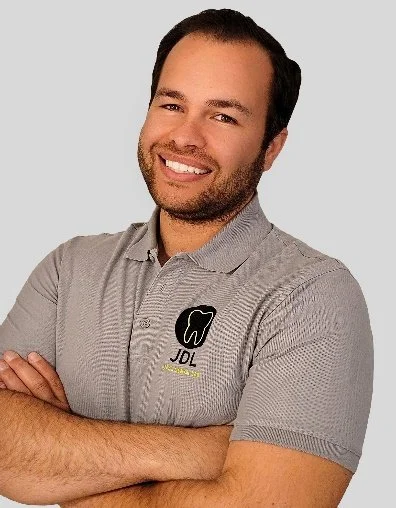A man with dark hair and a beard smiling, wearing a gray polo shirt with a tooth logo and JB Dental written on it, arms crossed.