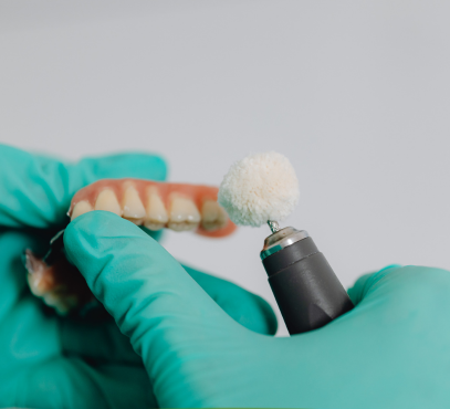 Close-up of a dental technician's hand wearing a teal glove, holding a dental prosthetic with a polishing tool.