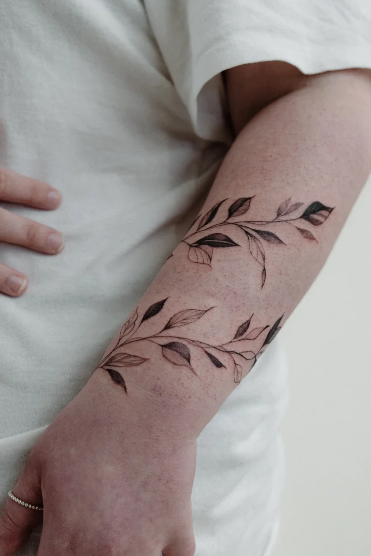 Fine line leaf vine tattoo wrapping around a forearm in two bands, by Tara Rose