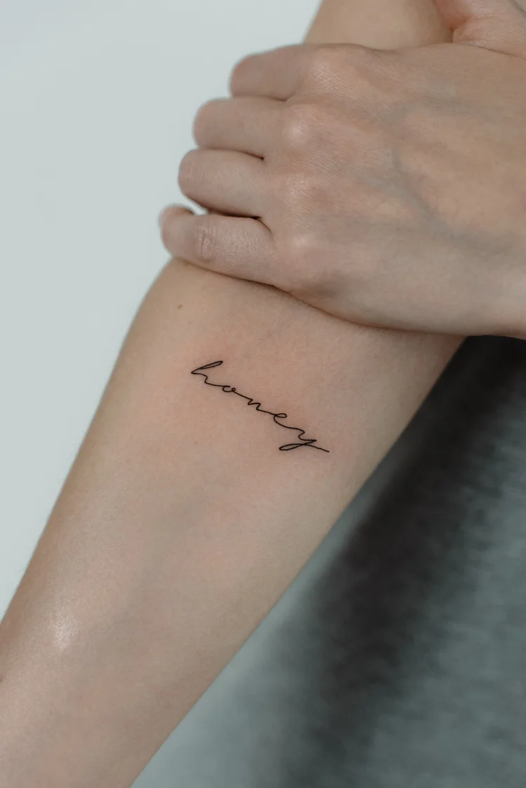 Fine line script tattoo reading honey on the forearm