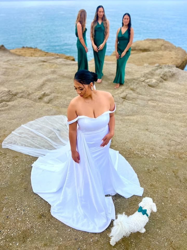 Bride in a bespoke off-shoulder white wedding gown with cathedral train, standing with bridesmaids in emerald green on a coastal clifftop