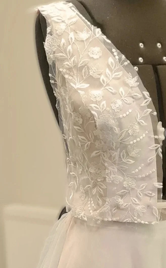 Close-up of a sustainably designed bespoke wedding gown bodice in progress, featuring intricate white ivory floral and leaf lace appliqué on sheer fabric, draped over a dress form with a soft tulle skirt