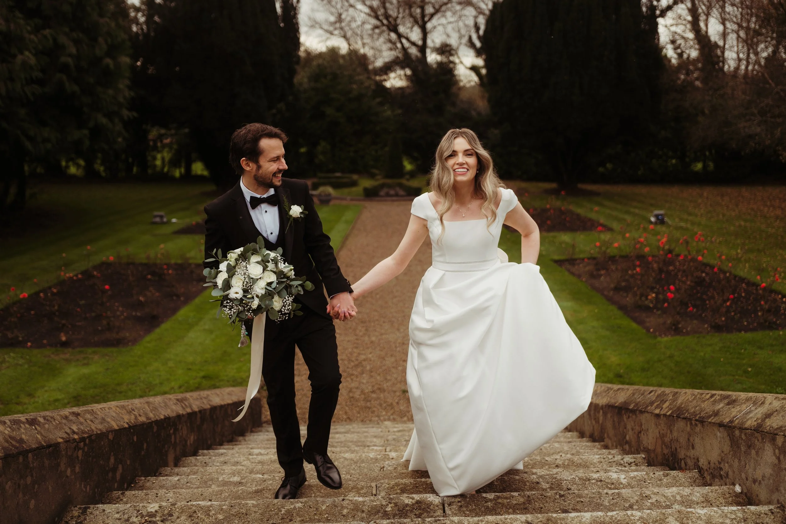 A bride and groom holding hands and walking up stone stairs in a garden, dressed in wedding attire. The bride is lifting her dress with one hand, and the groom is holding a bouquet of white flowers.