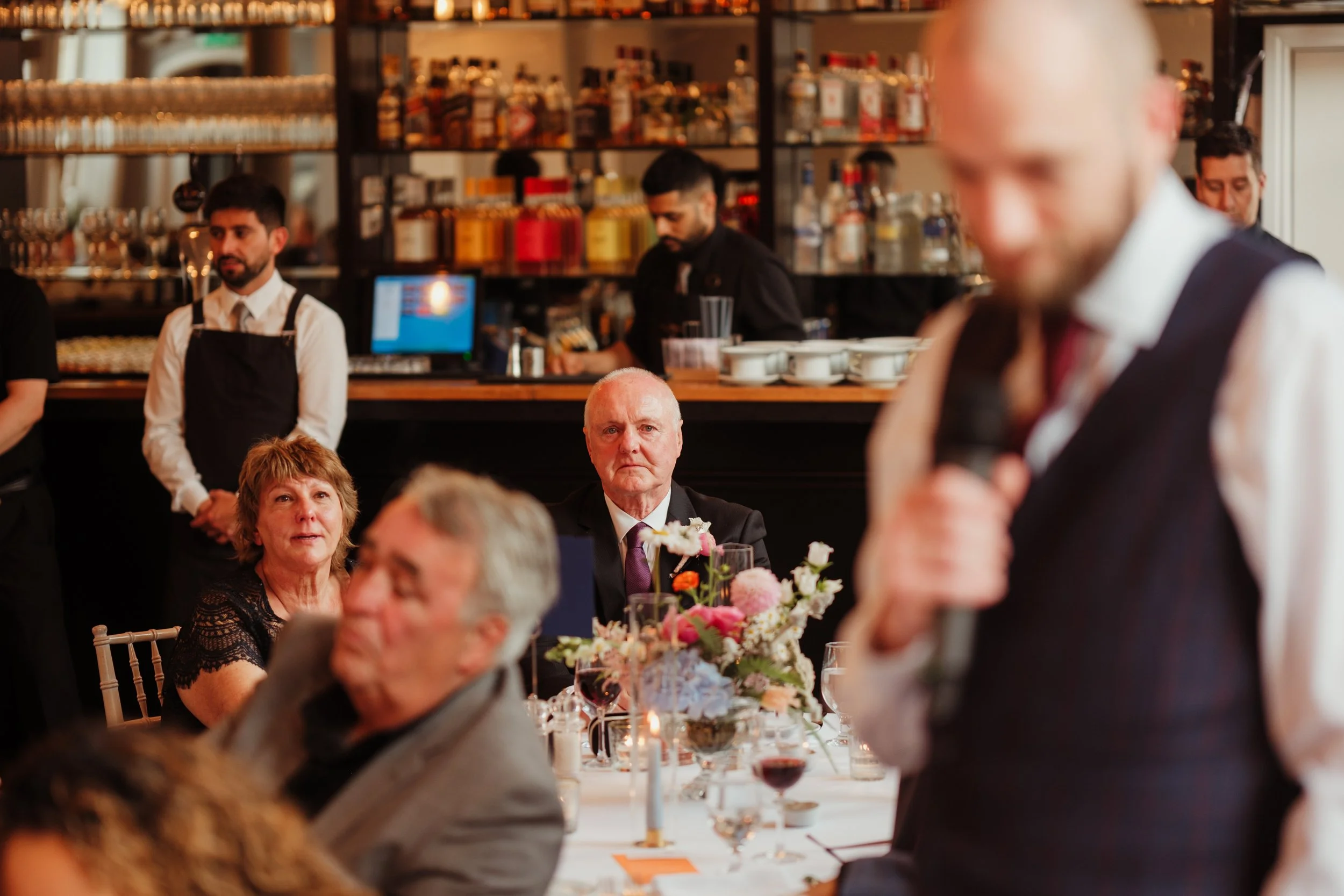 A man in formal attire giving a speech at a wedding reception, with guests seated at a decorated table, including an older man, a woman, and a man. The background shows a bar with bartenders and alcohol bottles.