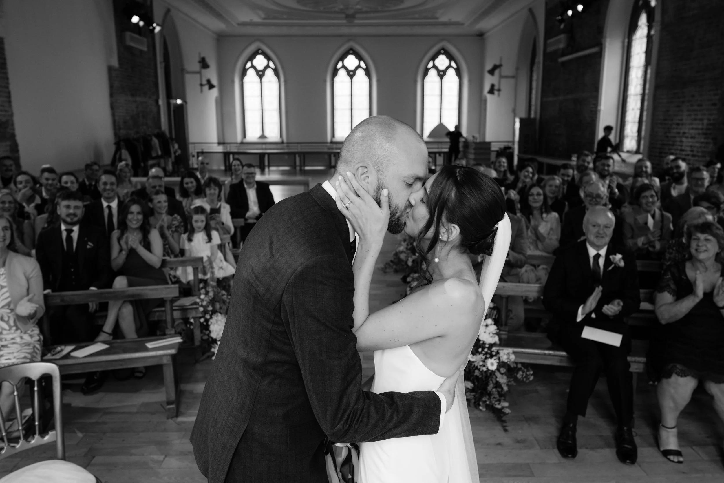 A black-and-white photo of a wedding ceremony showing a bride and groom kissing in front of guests in a chapel with large arched windows.