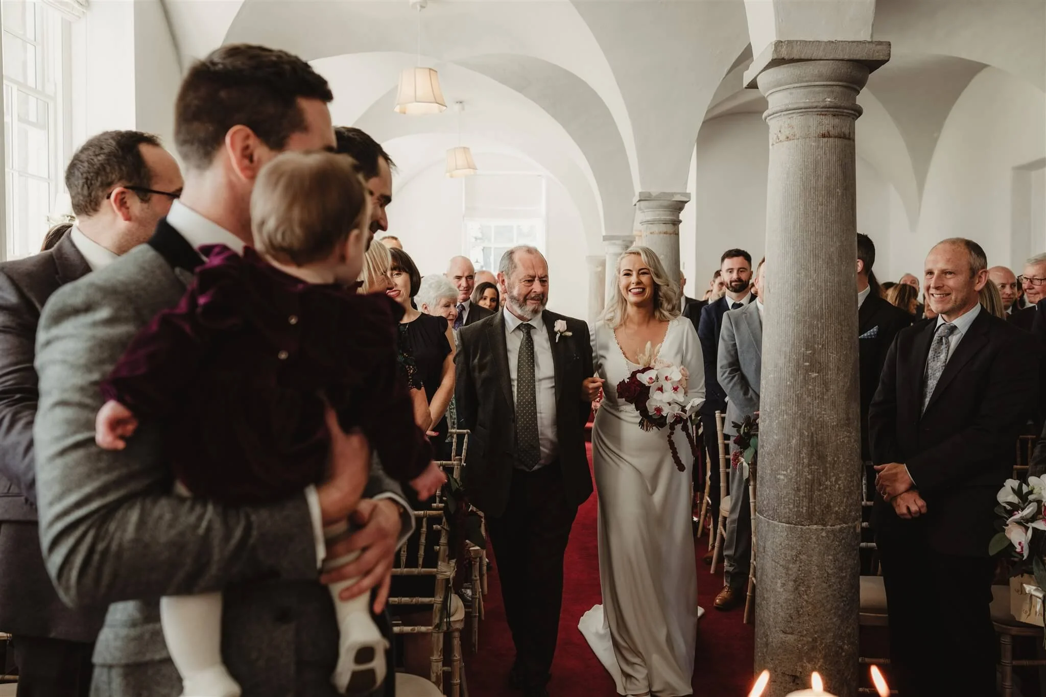 A bride walking down the aisle with her father while guests look on and smile in a church with white walls, arched ceilings, and stone columns.