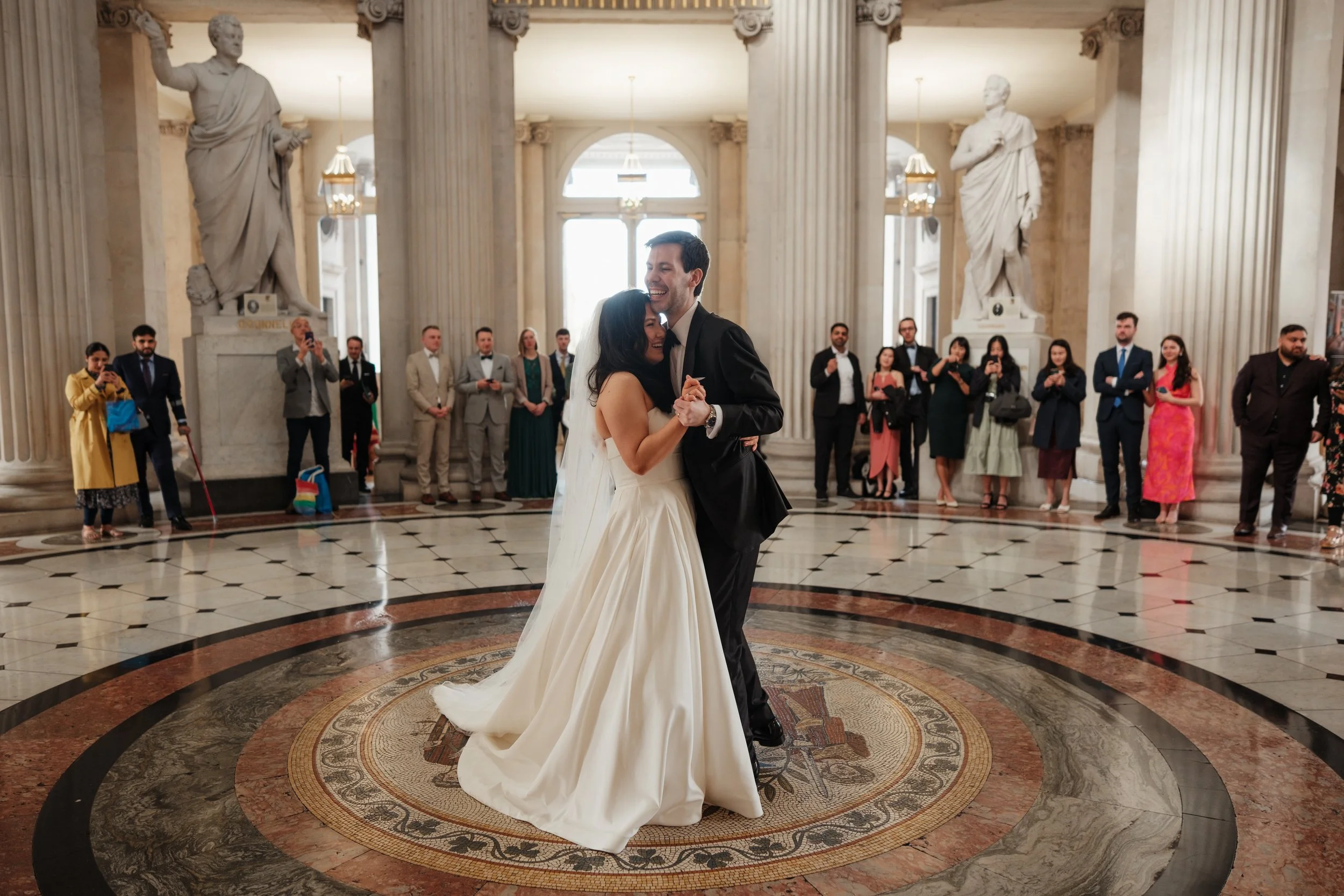 A bride and groom dancing happily in a grand hall with tall columns and statues, surrounded by guests watching and taking photos.