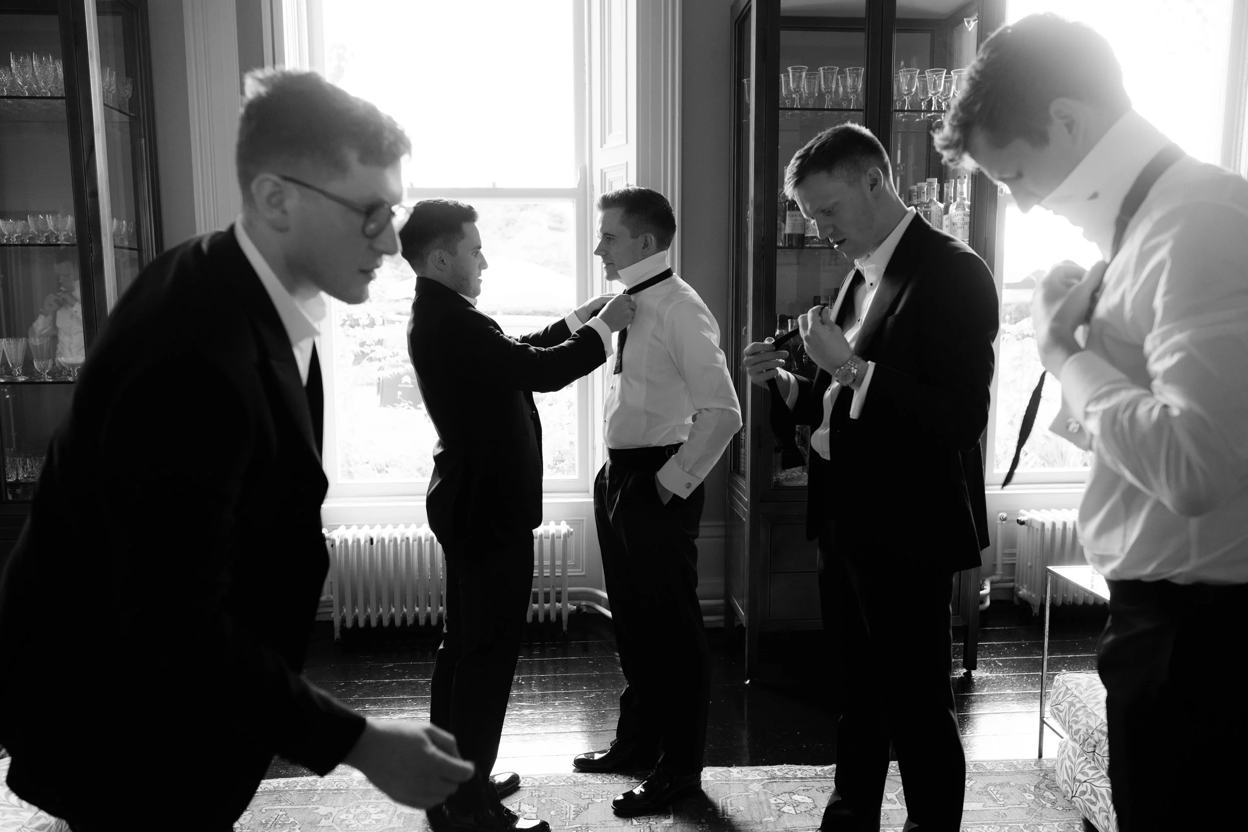 Black and white photo of five men in formal suits getting ready, with one adjusting the other's tie, in a room with large windows and glass cabinets.