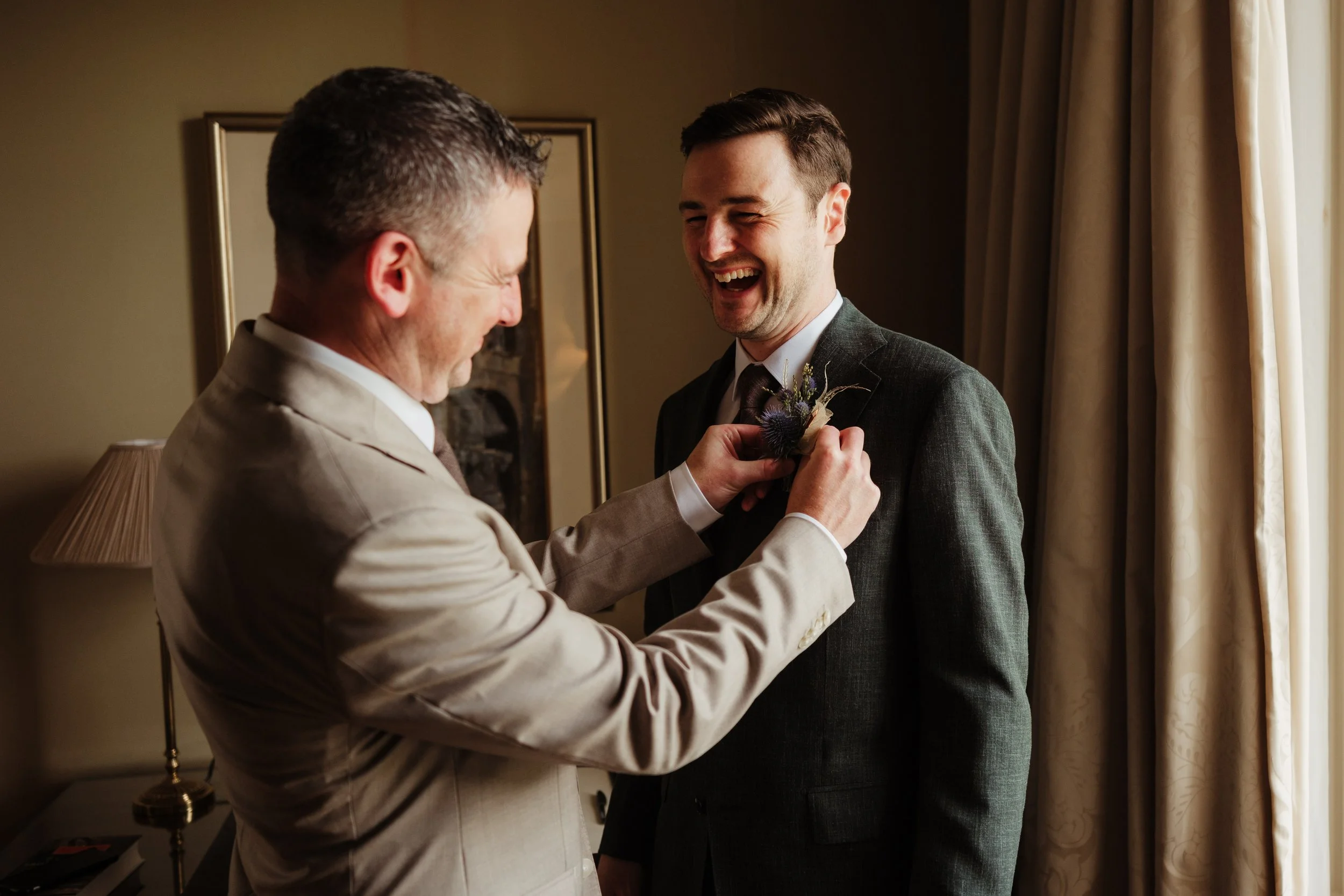 A man is helping another man pin a boutonniere on his suit jacket in a warmly lit room.