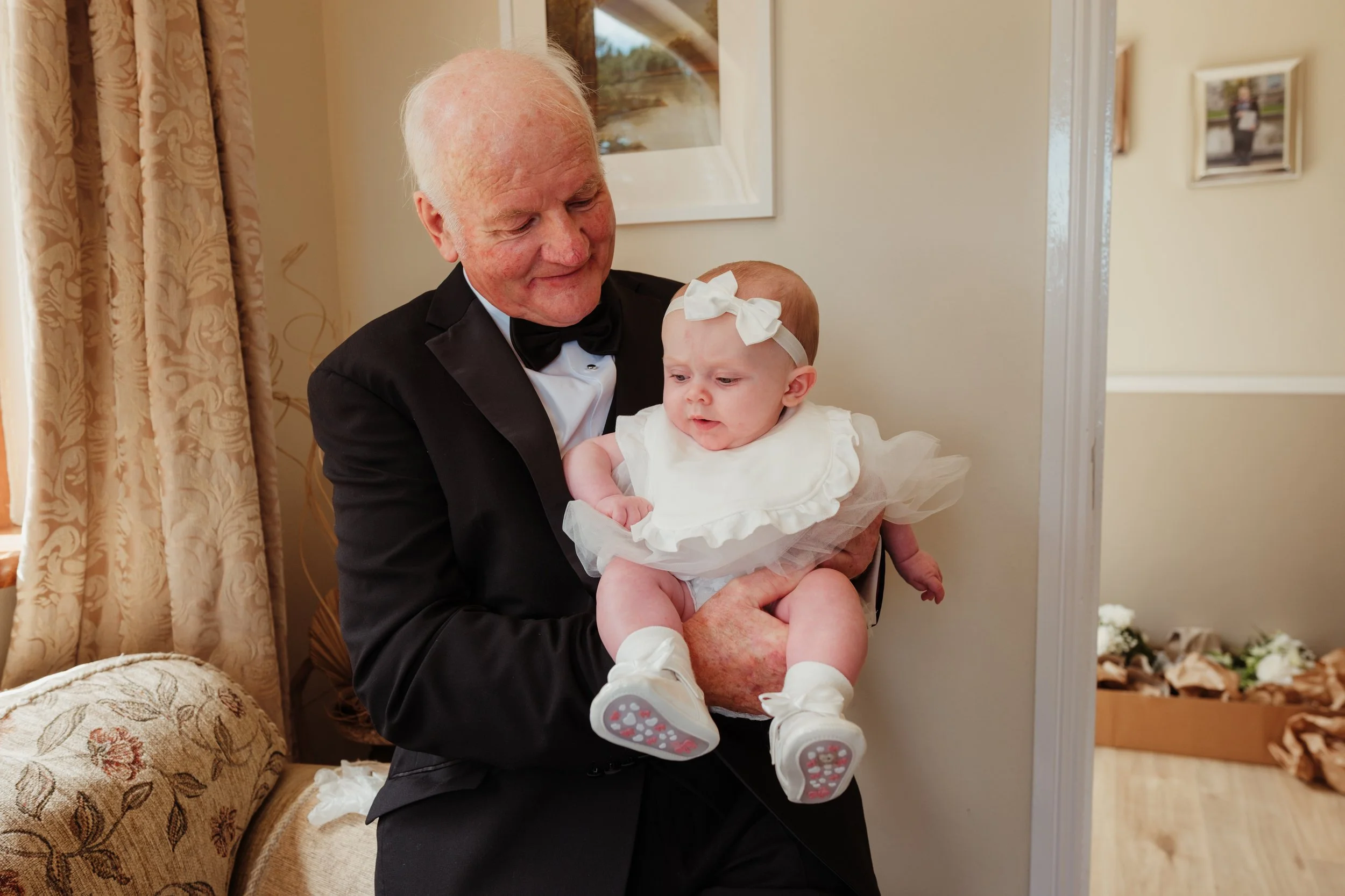An elderly man in a formal black tuxedo holding a baby girl dressed in a white dress and bow, inside a warmly lit room with beige walls, curtains, and framed photos.