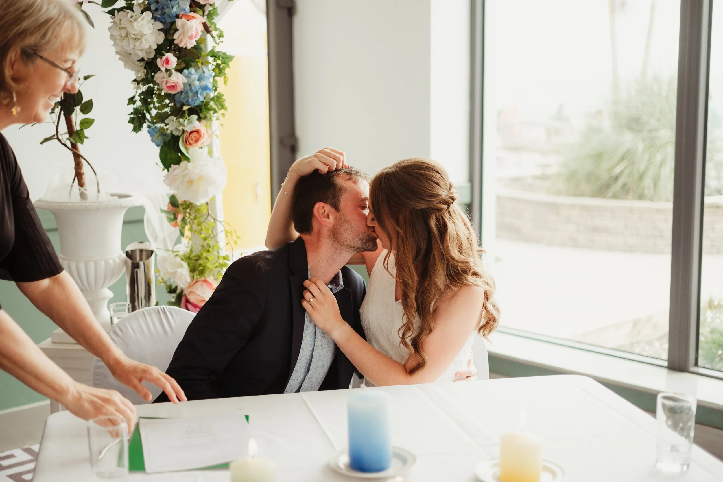 A couple shares a kiss during their wedding ceremony, seated at a table with wedding candles and floral arrangements, with an officiant standing nearby.