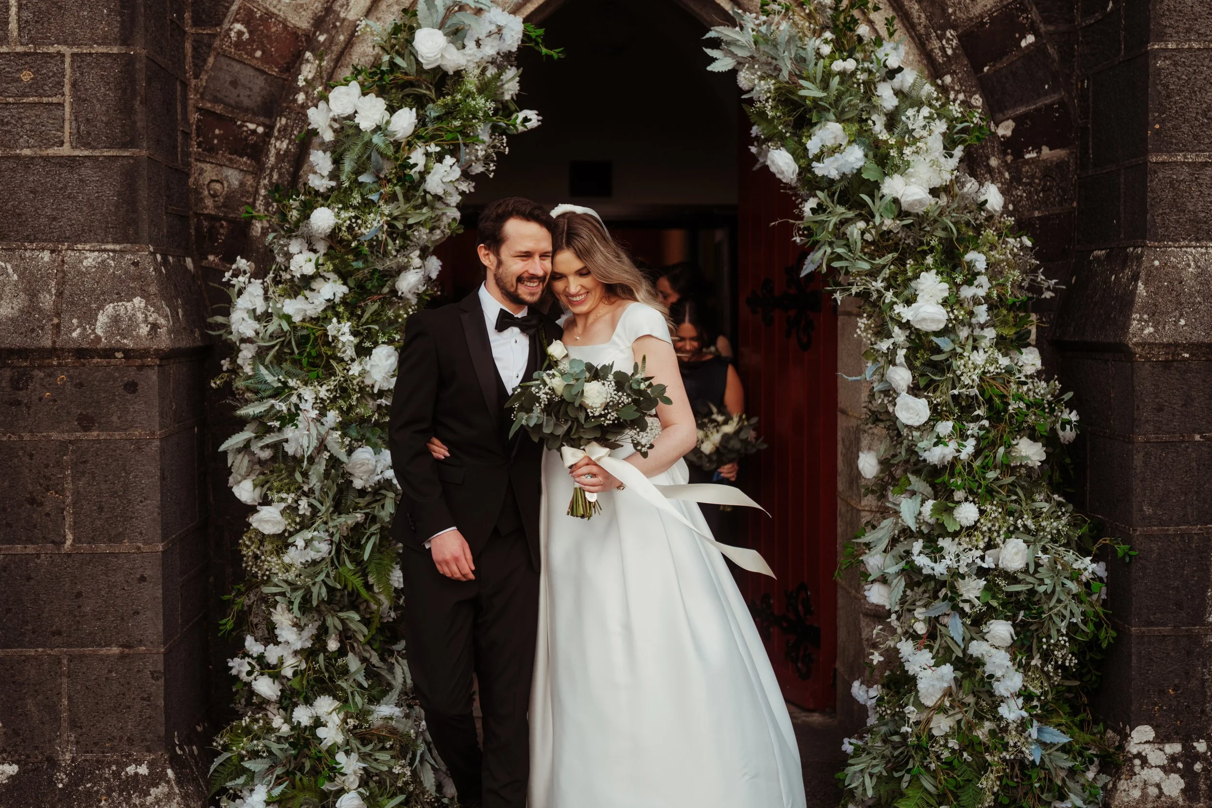 A bride and groom smiling, standing beneath a floral arch outside a stone building, with other bridesmaids in the background.