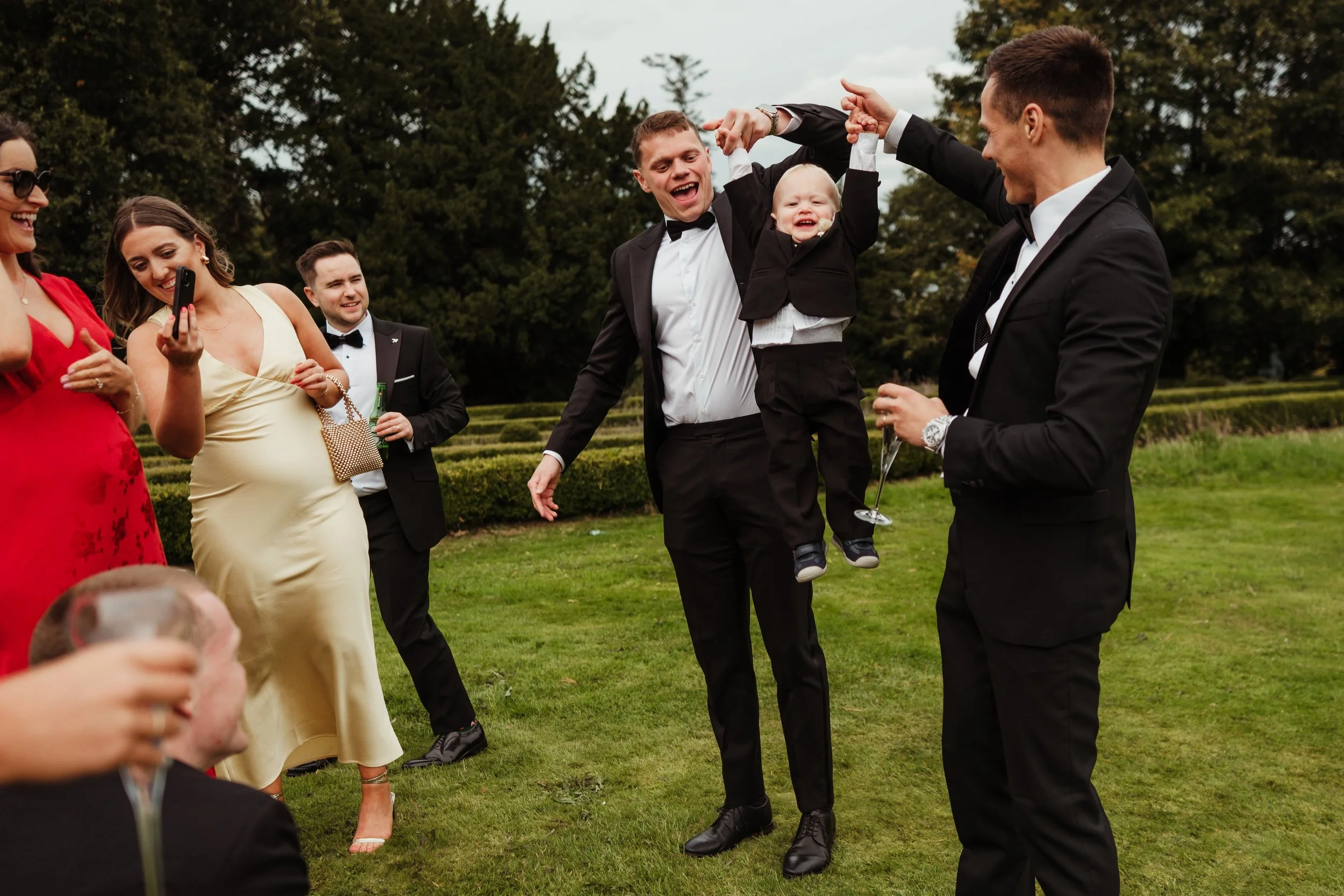 Group of people in formal attire celebrating outdoors, with a young boy lifting another child in a suit, surrounded by smiling adults.