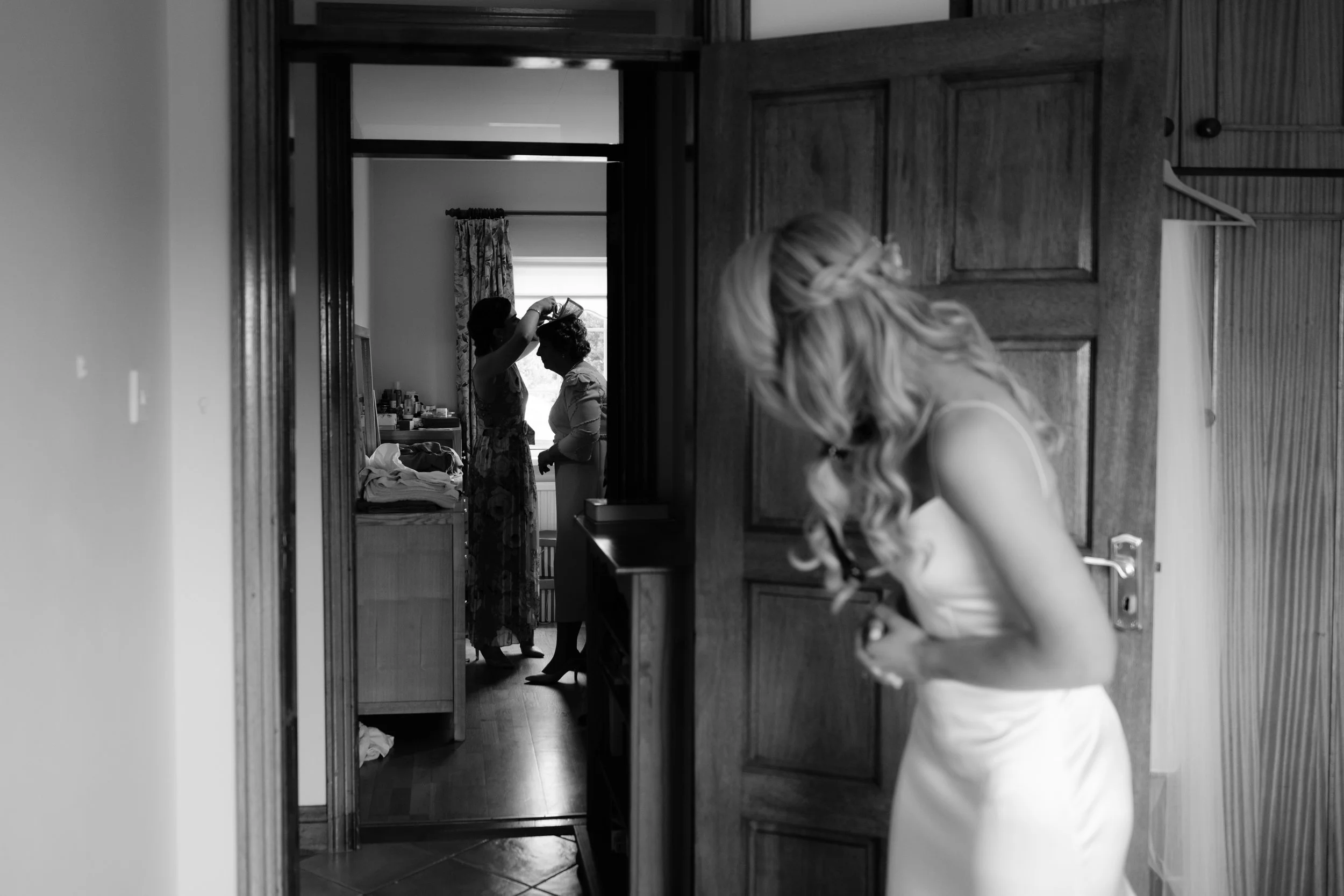 Bridal preparation scene in black and white, with one woman in the foreground adjusting her dress and two other women in the background getting ready, one helping the other with her hair.