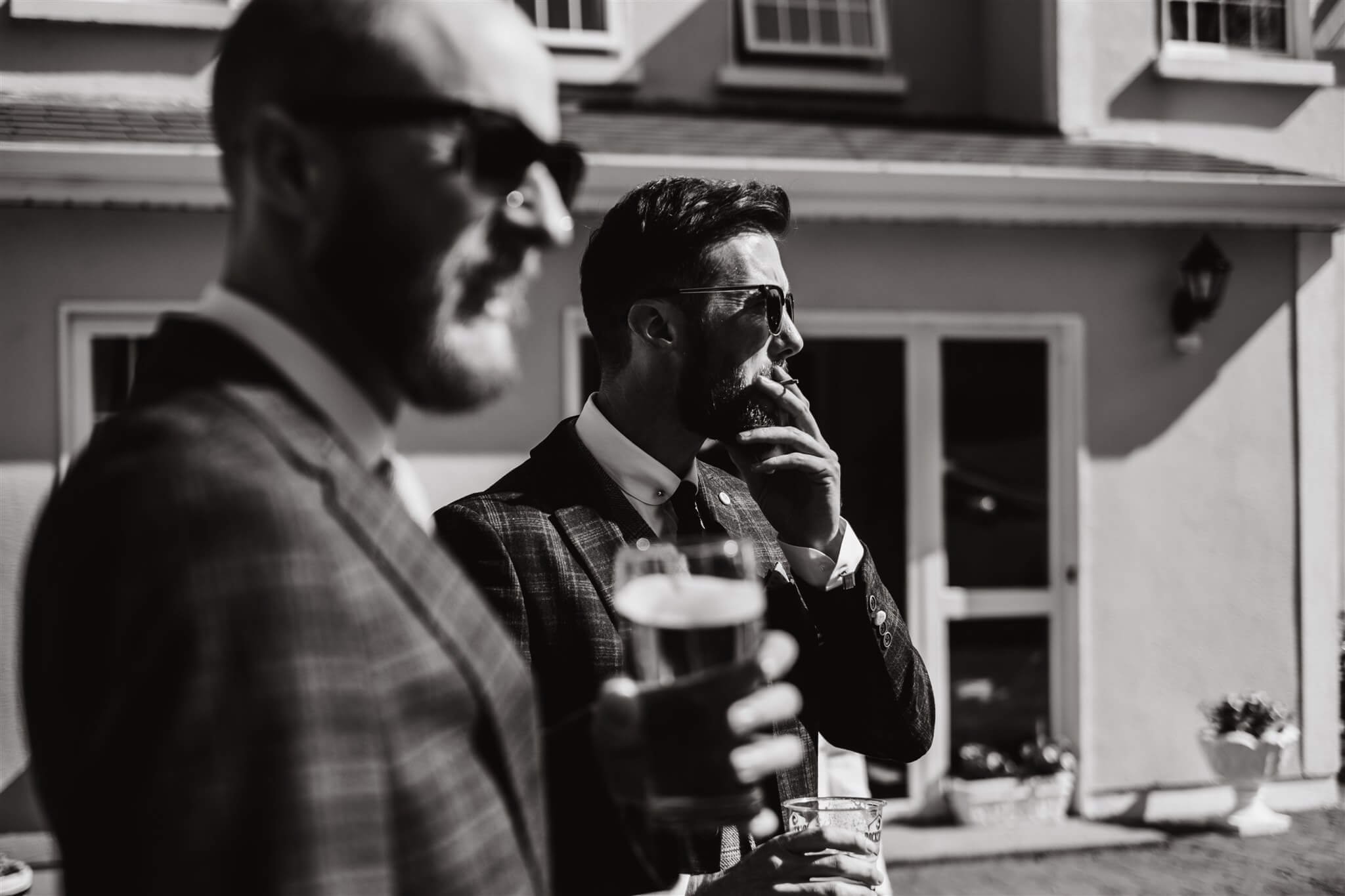 Two men in suits, one holding a glass of beer, standing outside in front of a house. The man on the right is wearing sunglasses and smoking a cigarette. Black and white photo.