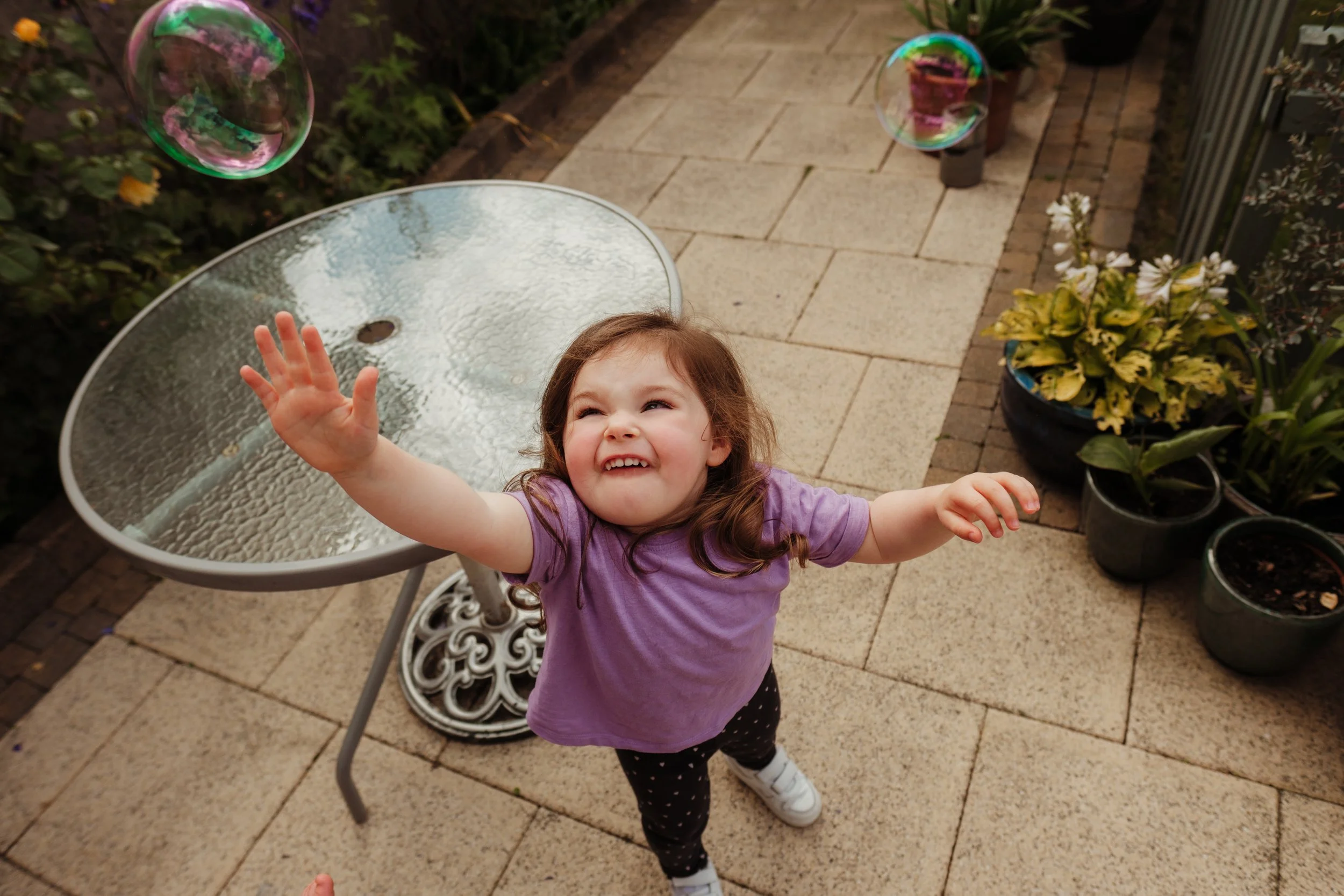 A young girl with brown hair reaching up towards floating soap bubbles in an outdoor patio setting.