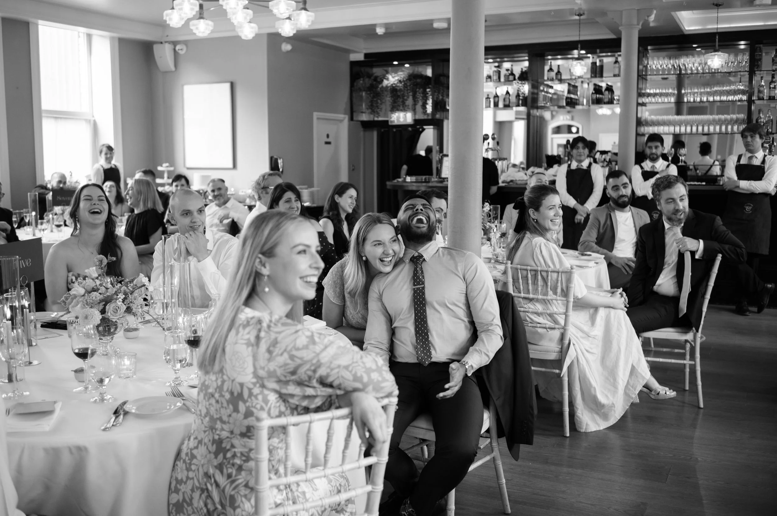 Group of people sitting at a banquet table, laughing and enjoying a social event in a decorated venue with waitstaff in the background.