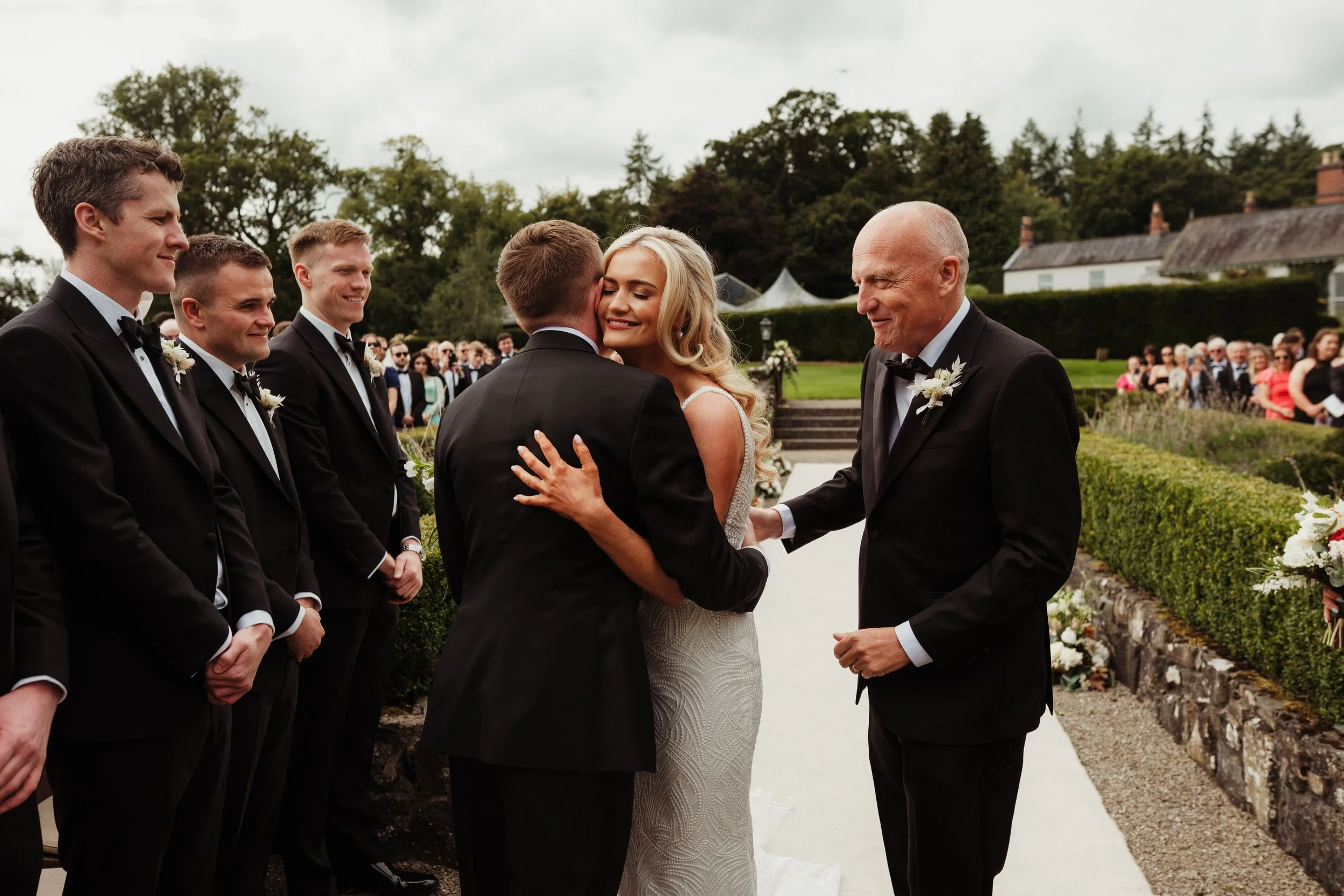 A bride and groom embrace during their wedding ceremony outdoors, with bridesmaids and groomsmen standing nearby, and guests in the background.