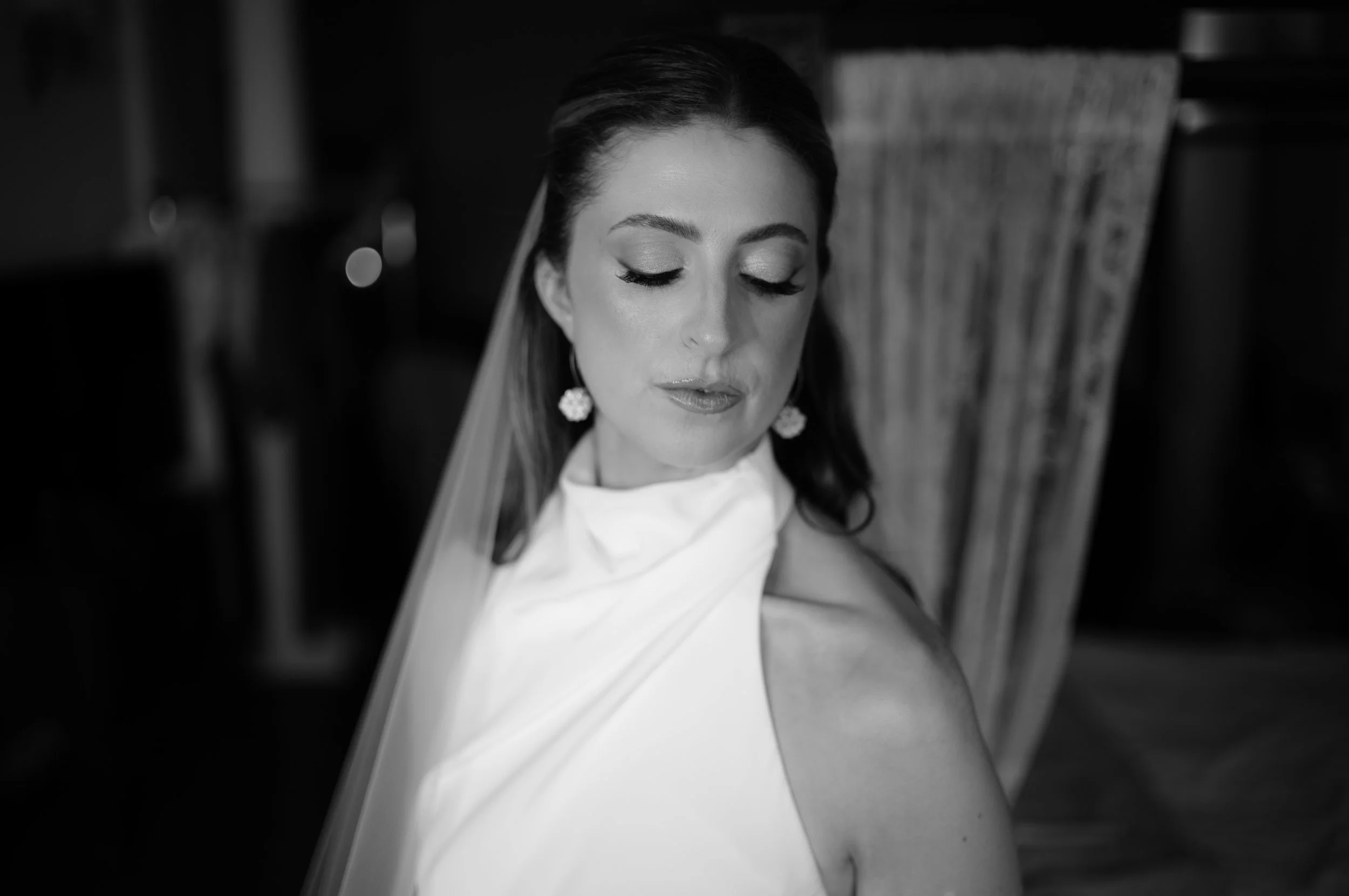 A black and white photograph of a woman with her eyes closed, wearing elegant earrings, a sleeveless dress, and a veil, seated in a high-backed chair.