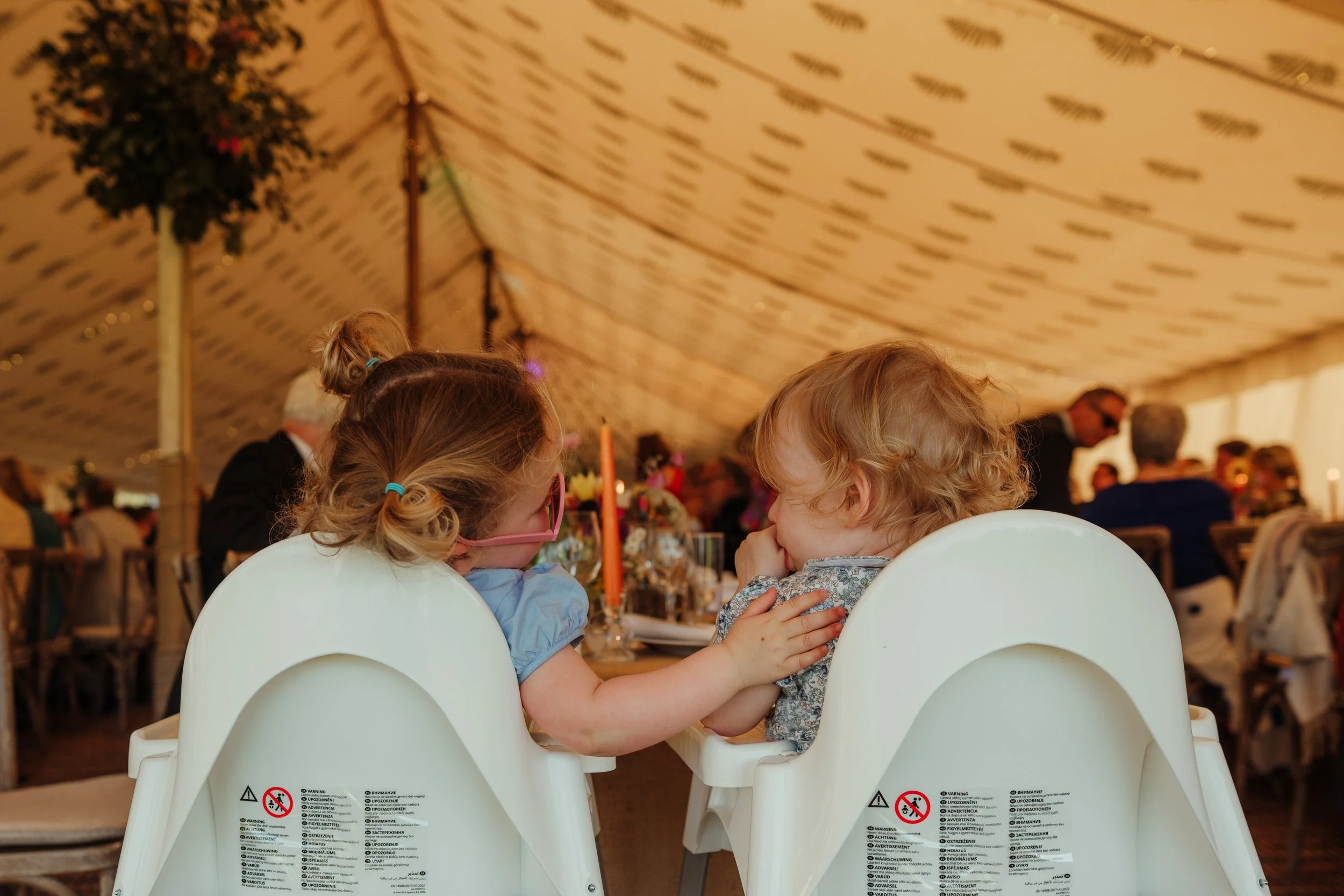 Two young children sitting in white high chairs at a table inside a decorated event tent, engaged in conversation.