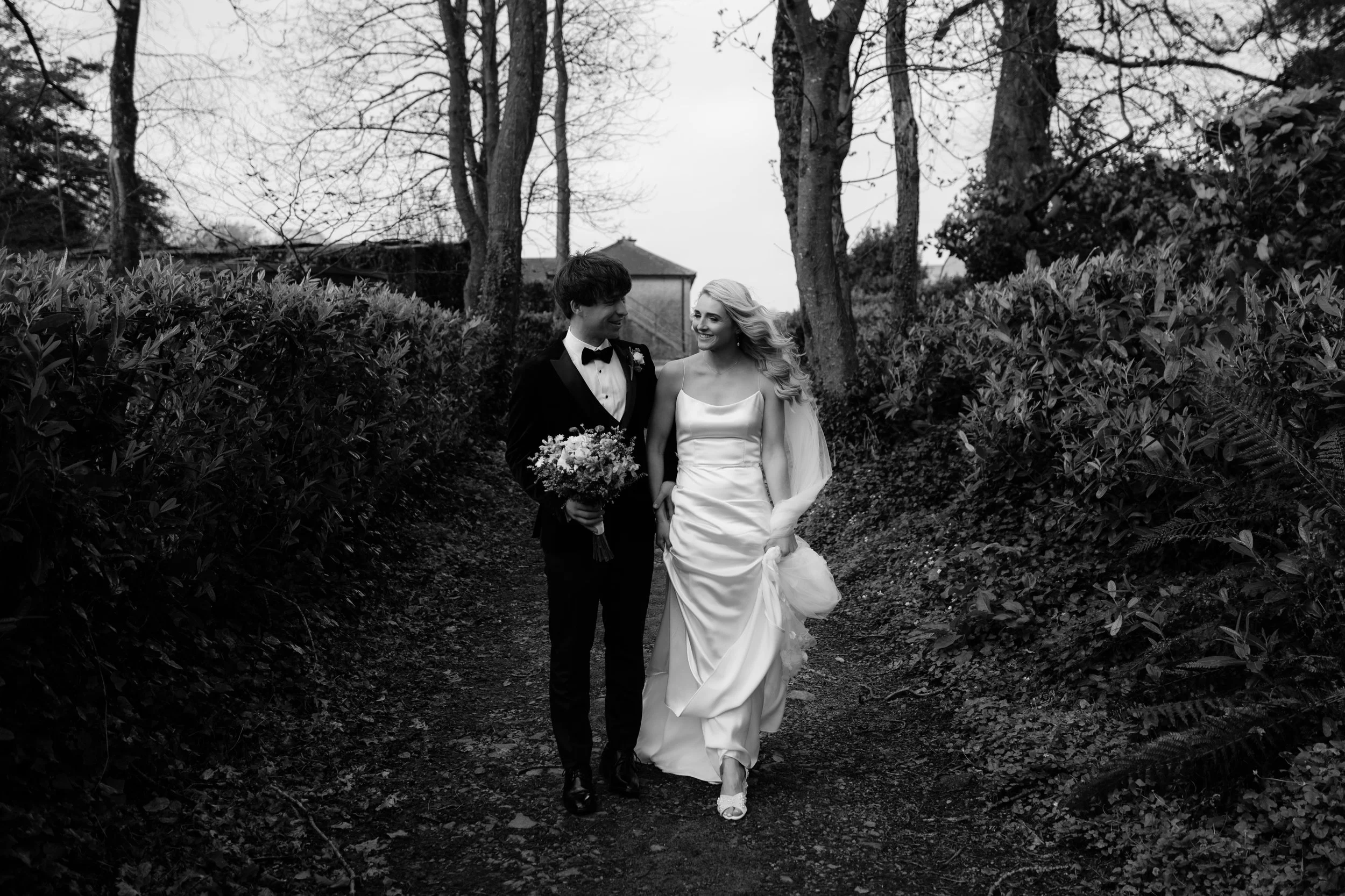 Black and white photo of a smiling bride and groom walking on a dirt path through a wooded area, surrounded by tall bushes and trees, with a house visible in the background.