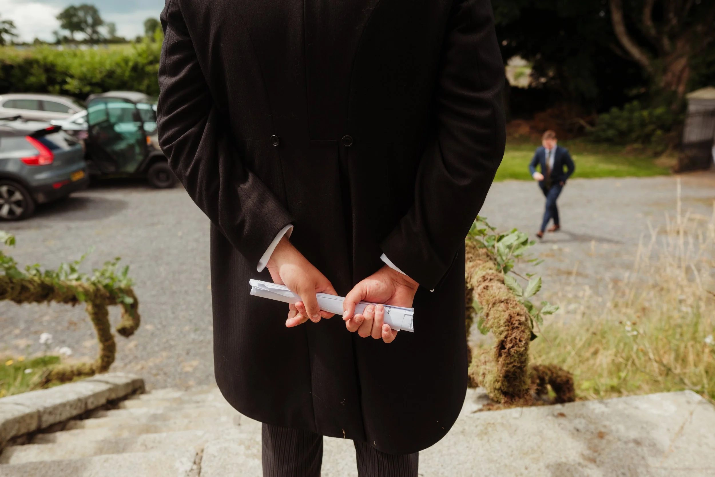 A person in a black coat holding rolled-up documents behind their back standing on outdoor steps, with another person in a suit walking in the background on a gravel area.