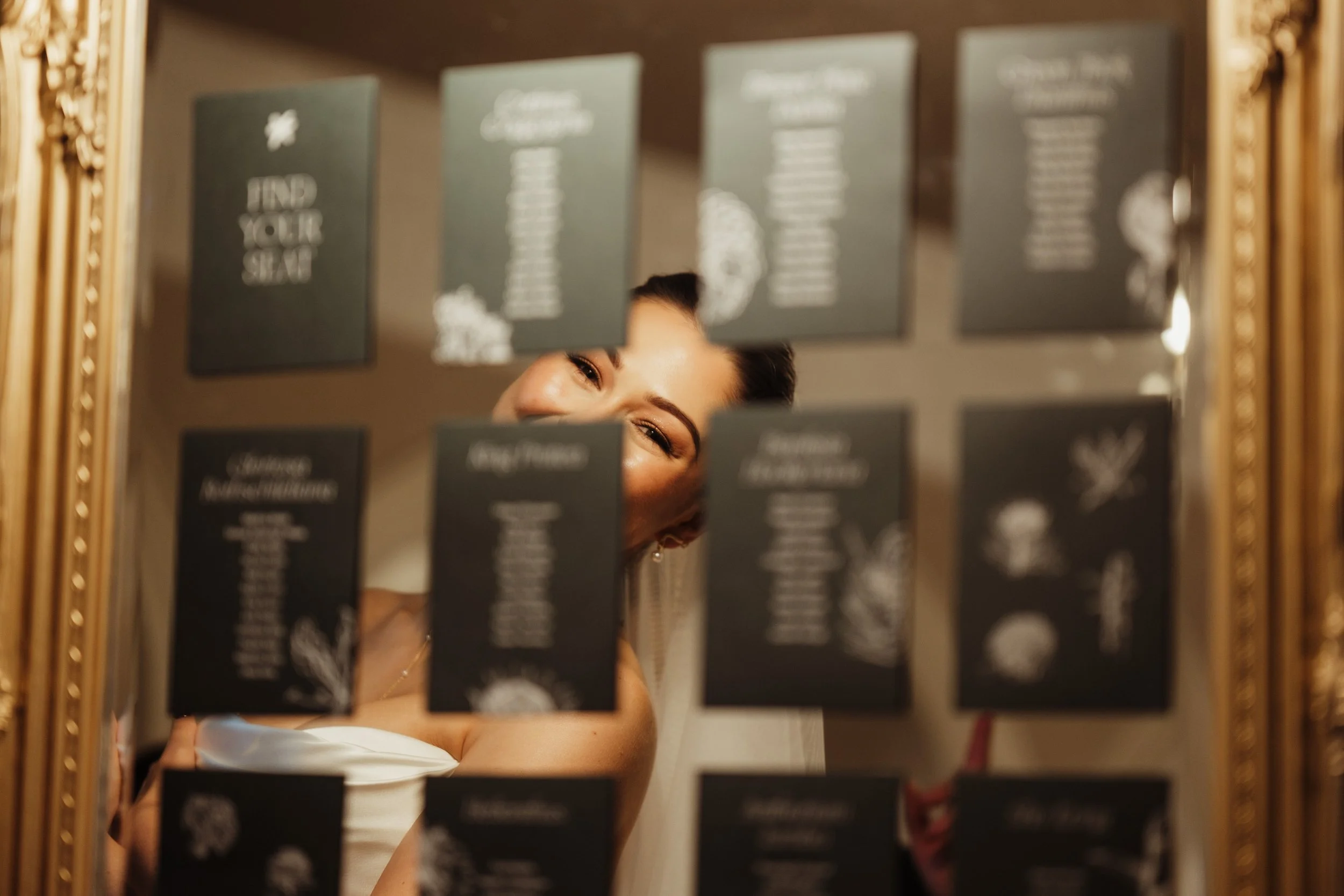 A woman smiling and looking at the mirror, partially obscured by hanging menu cards in a gold frame.