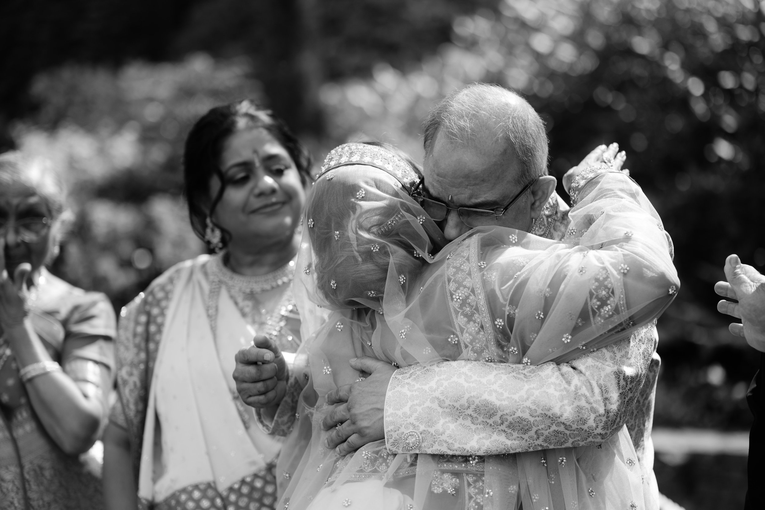 A black and white photo of an emotional family gathering, with one woman hugging an elderly man, while others look on, dressed in traditional Indian attire.