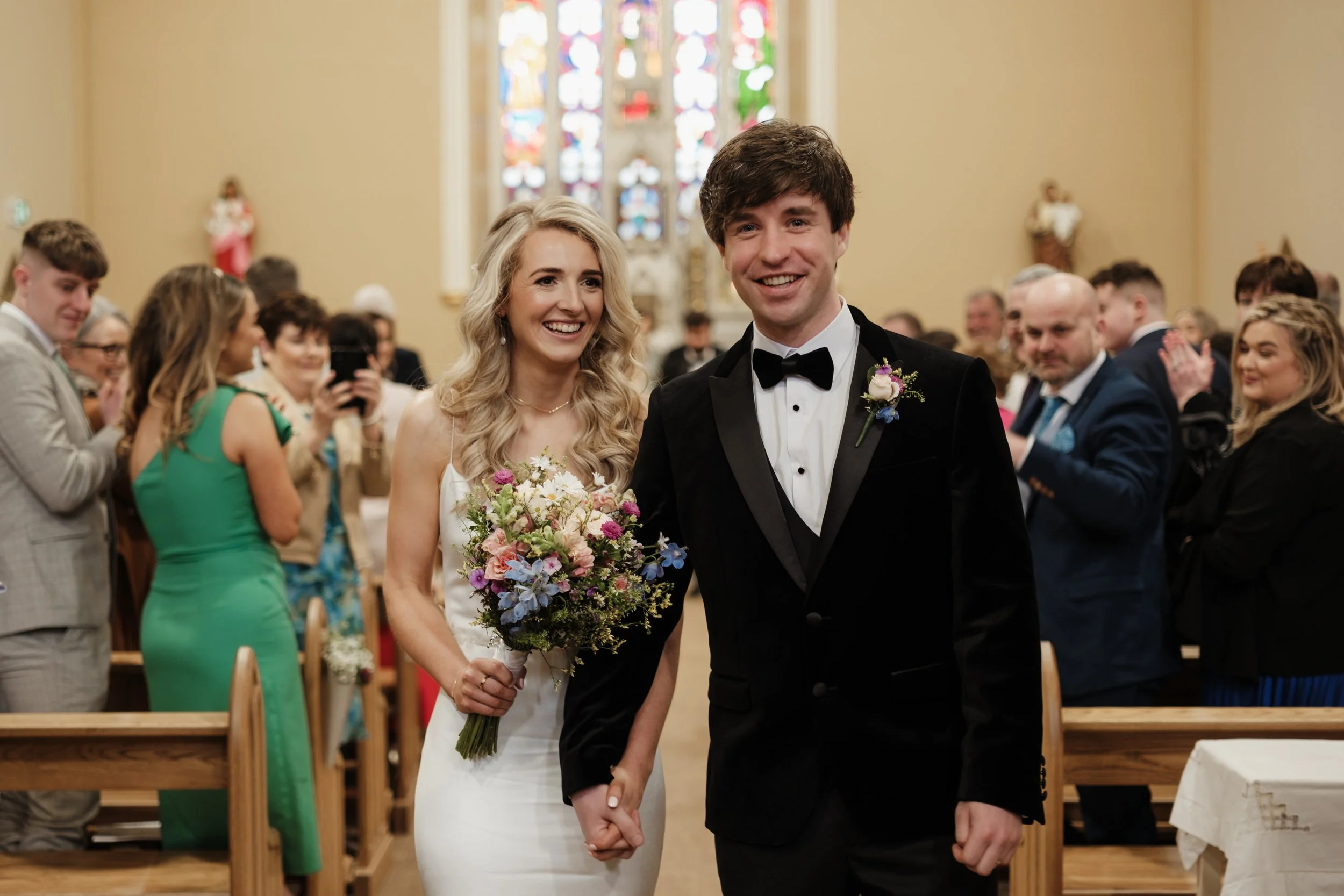 A bride and groom holding hands and smiling during their wedding ceremony, surrounded by guests inside a church with stained glass windows.