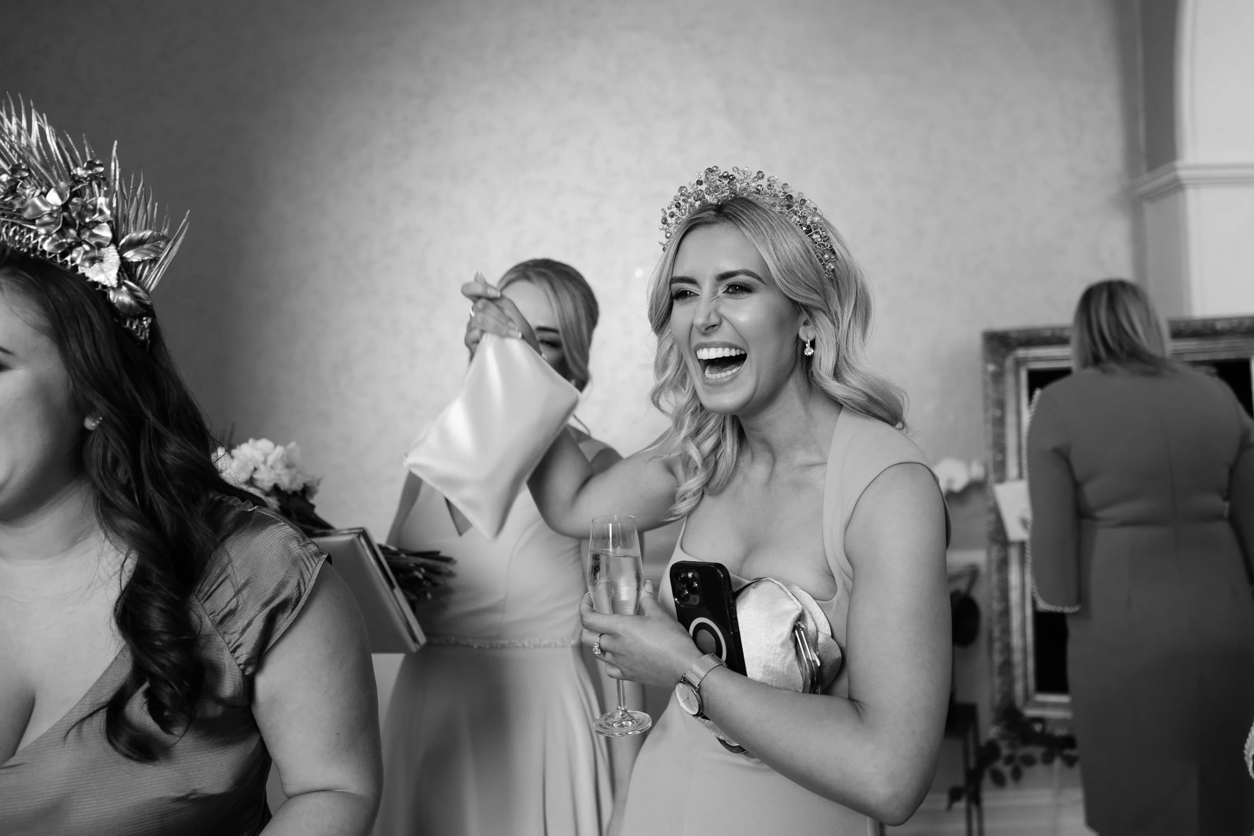 A woman at a celebration, wearing a crown, holding a champagne glass, laughing, and holding a clutch and phone in a decorated indoor setting.