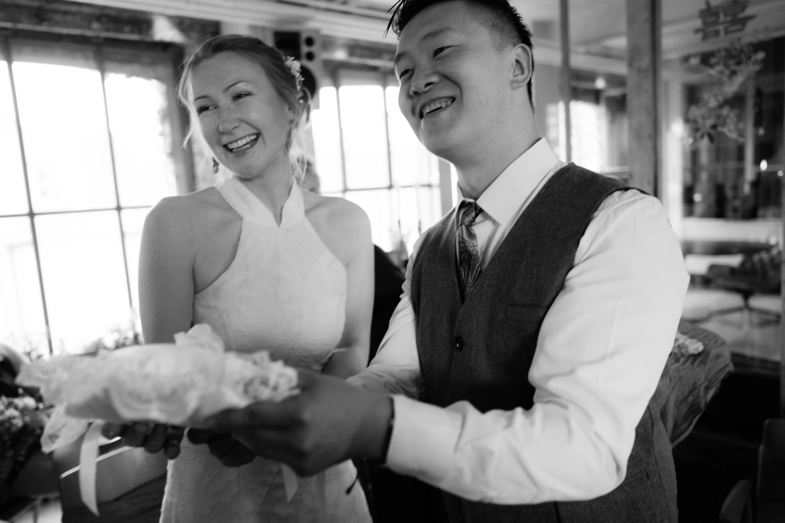 A bride and groom smiling during their wedding celebration, with the groom holding a tray of a wrapped gift in a festive, decorated indoor setting.