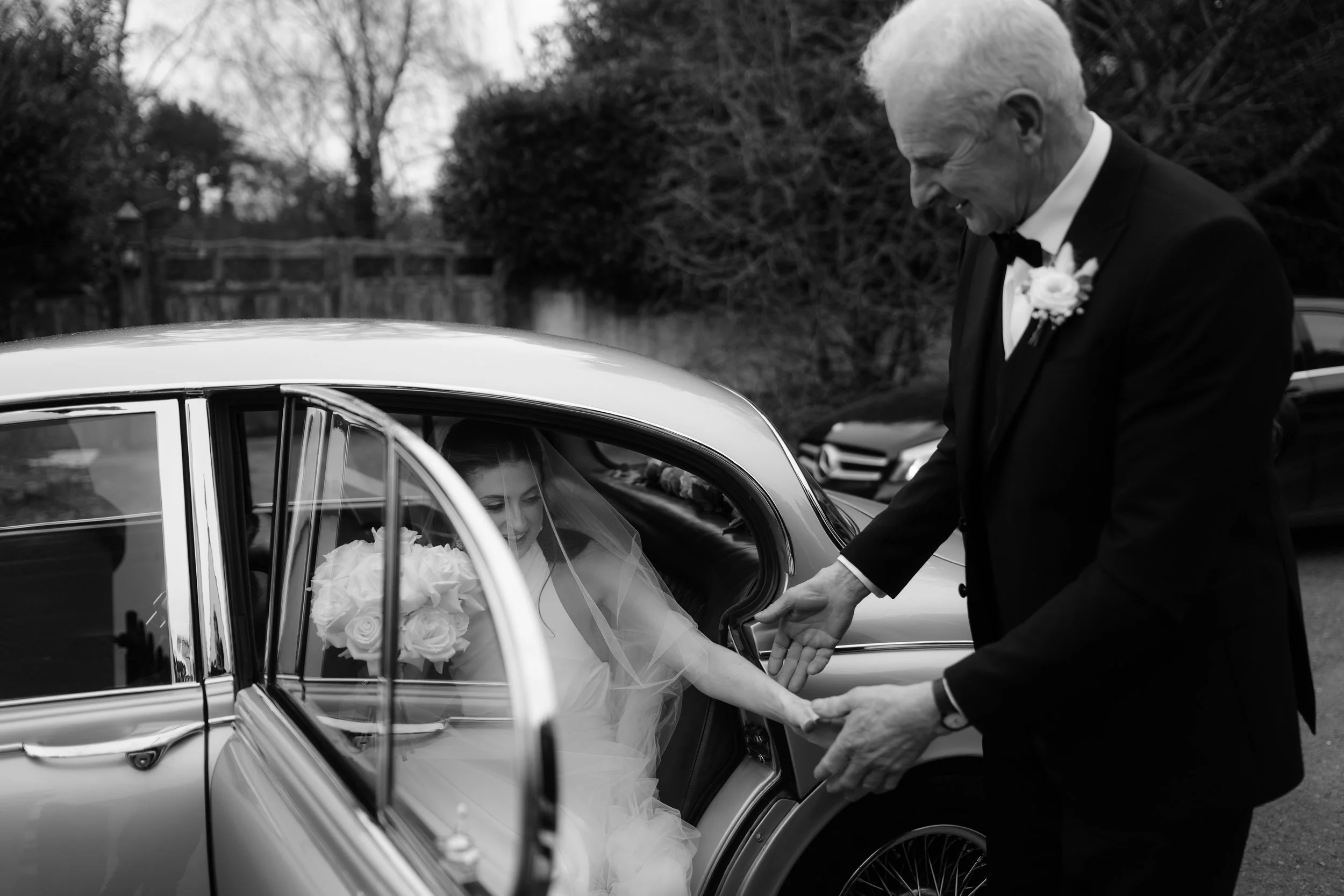A bride sitting in a vintage car holding a bouquet of roses, seen through the car window, as an older man, dressed in a tuxedo with a boutonniere, tenderly holds her hand outside the car on a cloudy day in a suburban neighborhood.