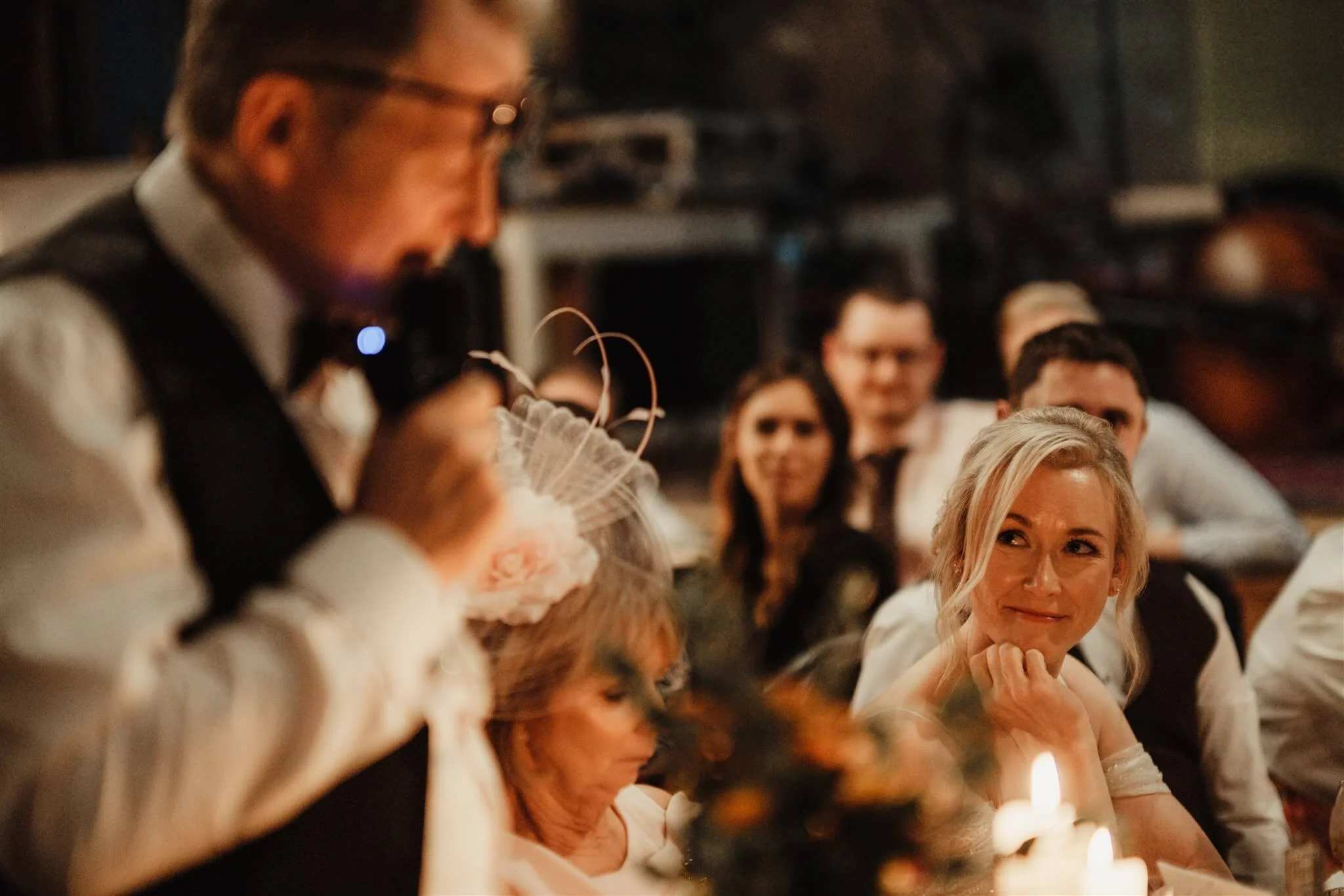 A man in a vest and glasses speaking at a wedding reception, with a woman in an off-shoulder dress smiling and resting her chin on her hand at the table, surrounded by guests.