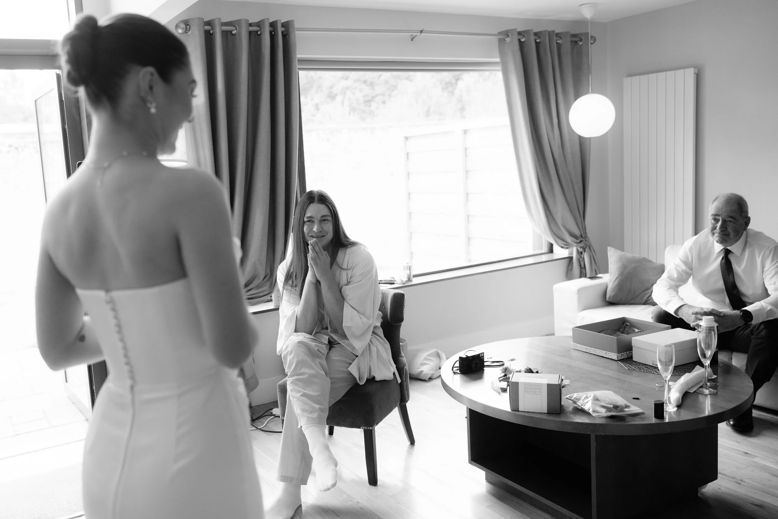 A woman in a wedding dress smiling and looking at two guests seated in a living room, one woman and one man. The guests appear emotional and happy, with the woman holding her hand to her face. The room has large windows with curtains, a coffee table 