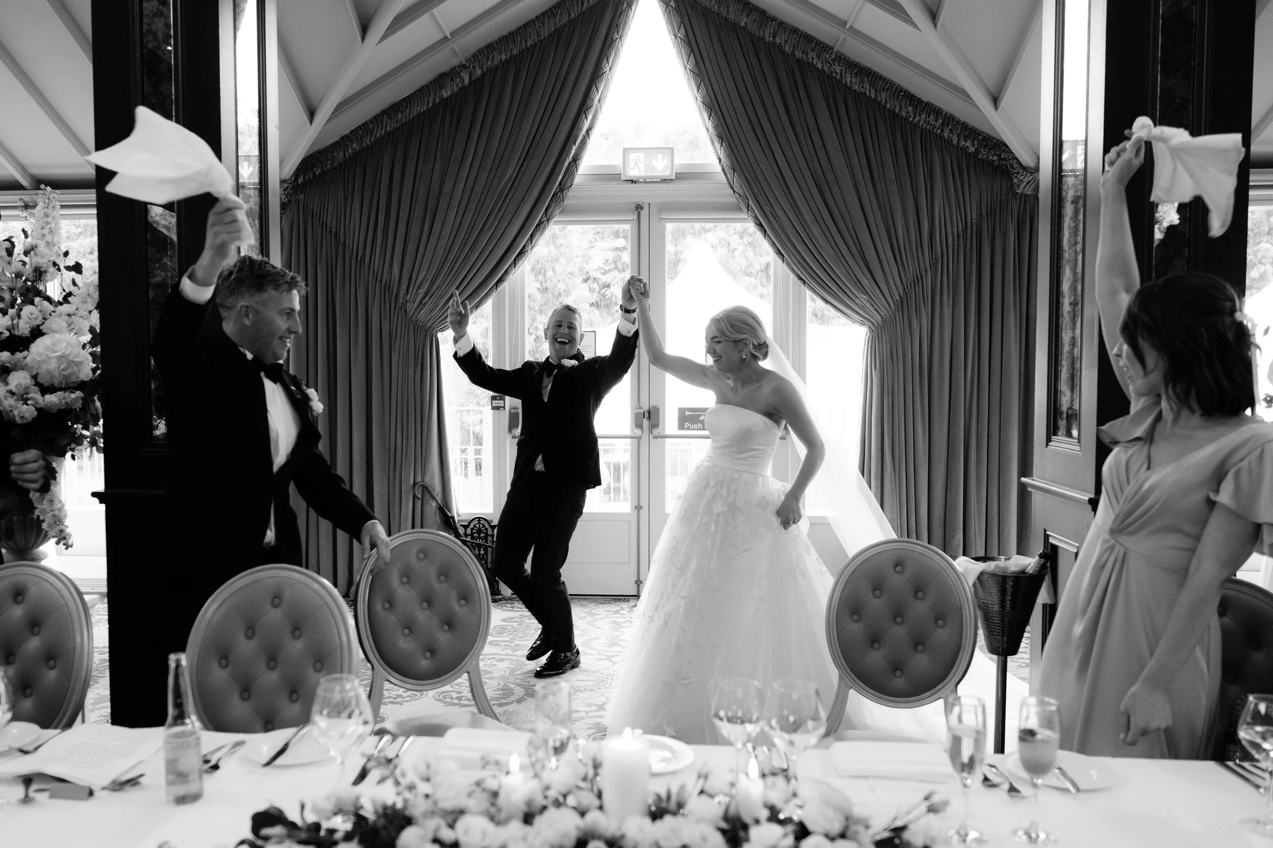 A black and white photo of a wedding celebration with a bride and groom holding hands and smiling while dancing, surrounded by friends in a decorated venue with large curtains and a table set for a meal.