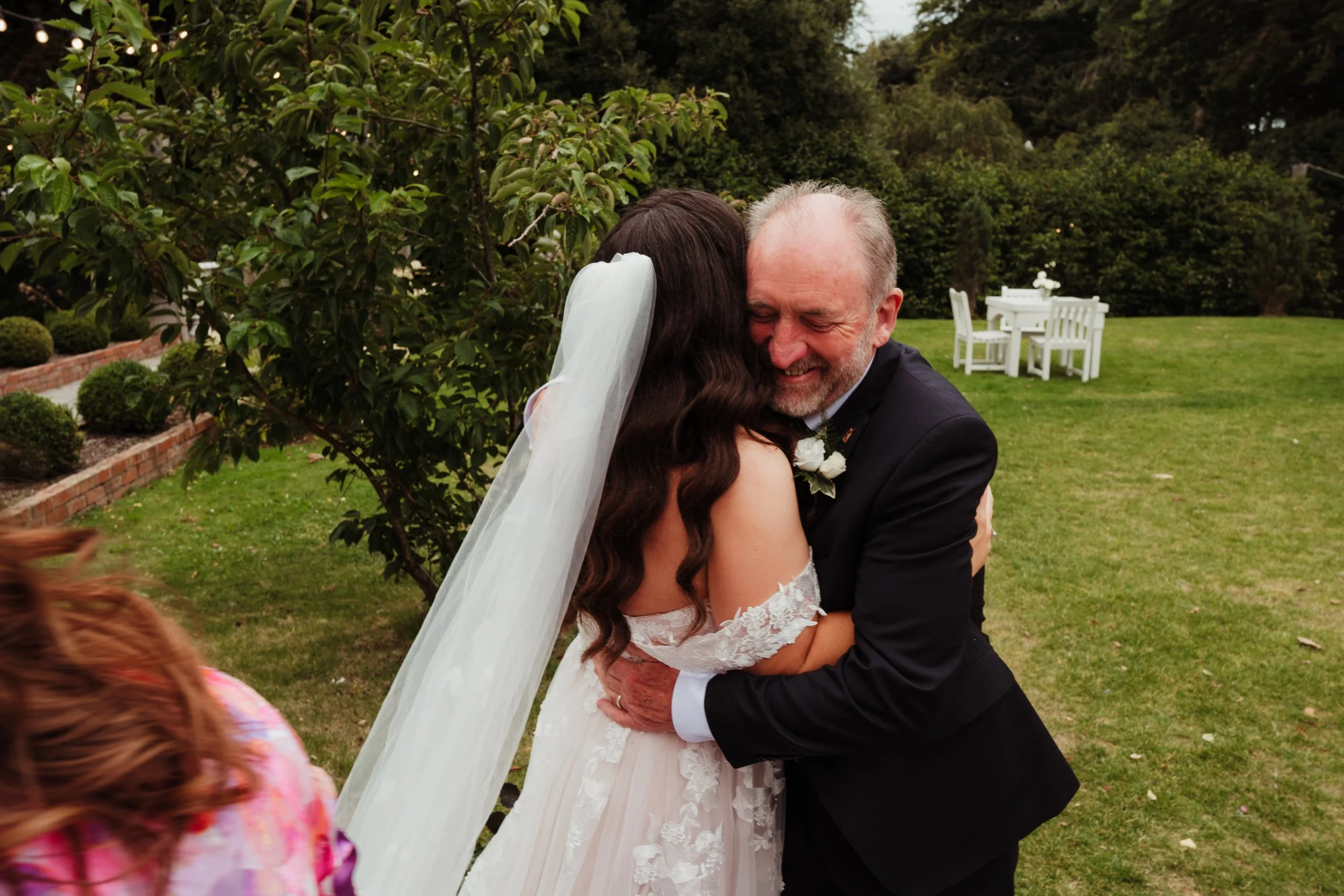 A bride and an older man sharing an emotional hug outdoors in a garden or park setting, with the bride wearing a lace off-shoulder wedding dress and a veil, and the man dressed in a dark suit with a boutonnière.