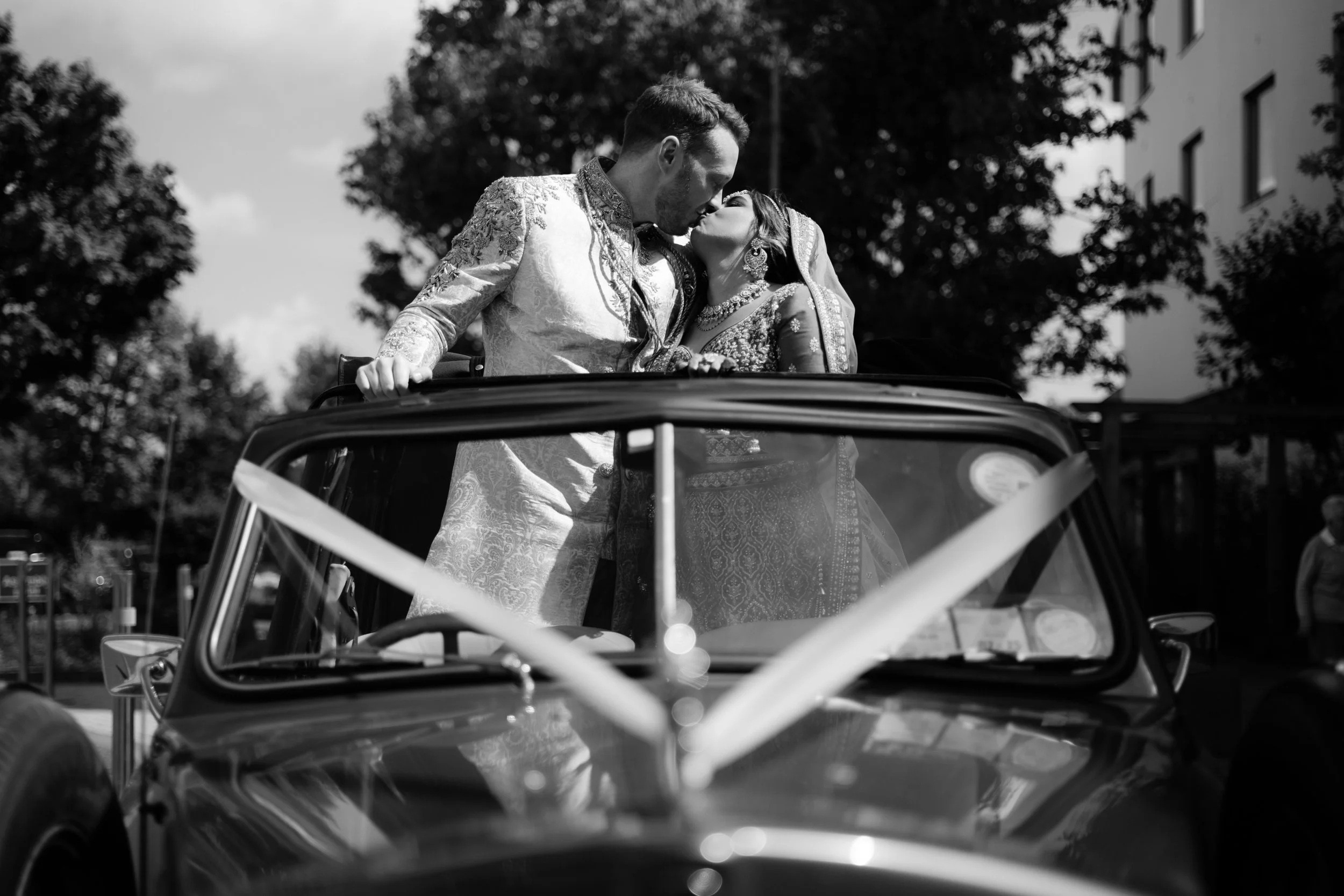 A couple dressed in traditional Indian wedding attire sharing a kiss while standing in a vintage car decorated with ribbons on the hood.