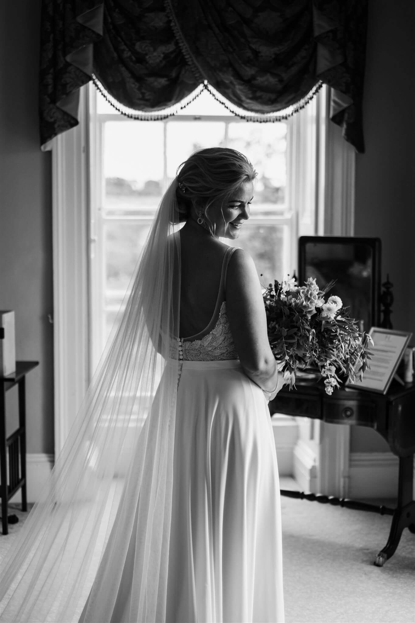 Black and white photo of a bride smiling, standing in front of a window with curtains, holding a bouquet of flowers, wearing a wedding dress and veil.