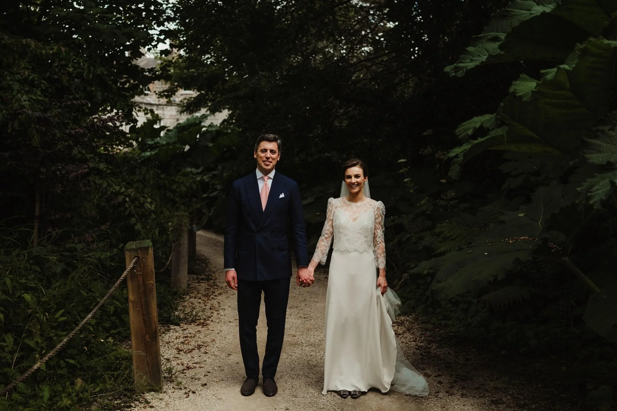 A bride and groom holding hands and walking along a wooded path on their wedding day, surrounded by lush green foliage.