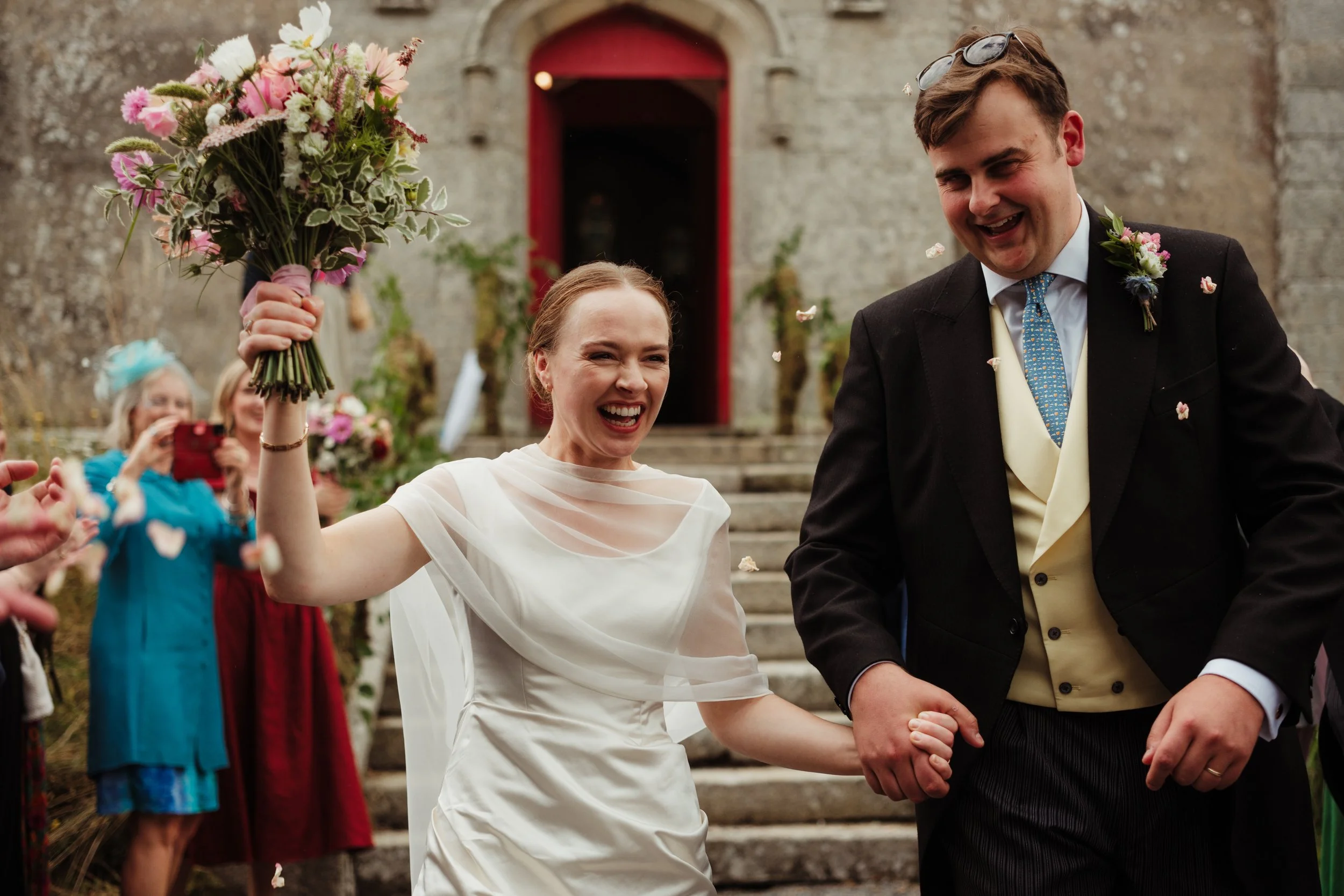 A happy bride and groom holding hands and celebrating outside a church after their wedding, with guests throwing flower petals in the background.