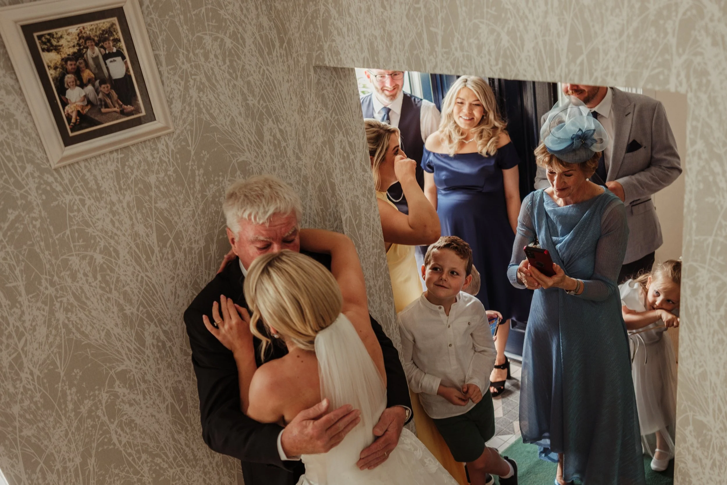 A wedding scene showing a bride and an older man hugging as guests look on, including a woman in a blue dress with a matching hat, a young boy smiling, and other women and men in formal attire.