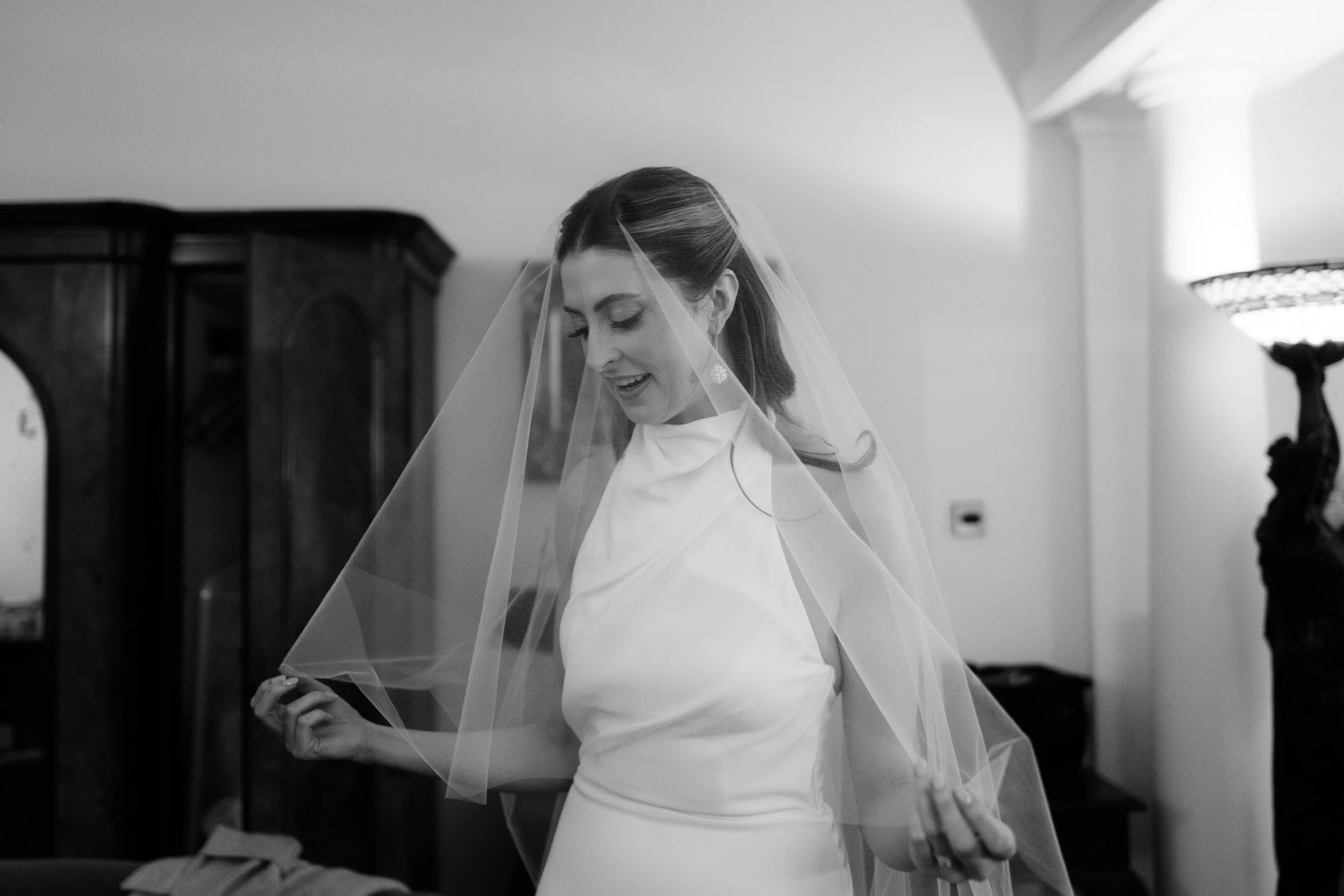 Black and white photo of a woman in a wedding dress with a veil, smiling and adjusting her veil indoors.