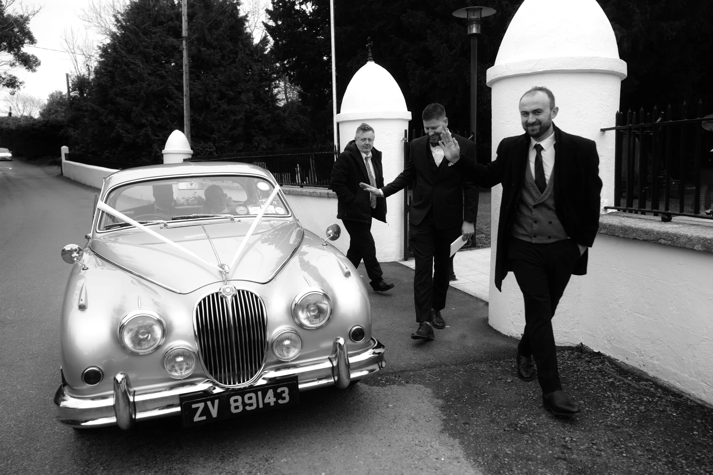 Three men in formal attire, two of whom are waving, standing by a vintage car decorated with ribbons near a white wall and a small building, in a street scene.