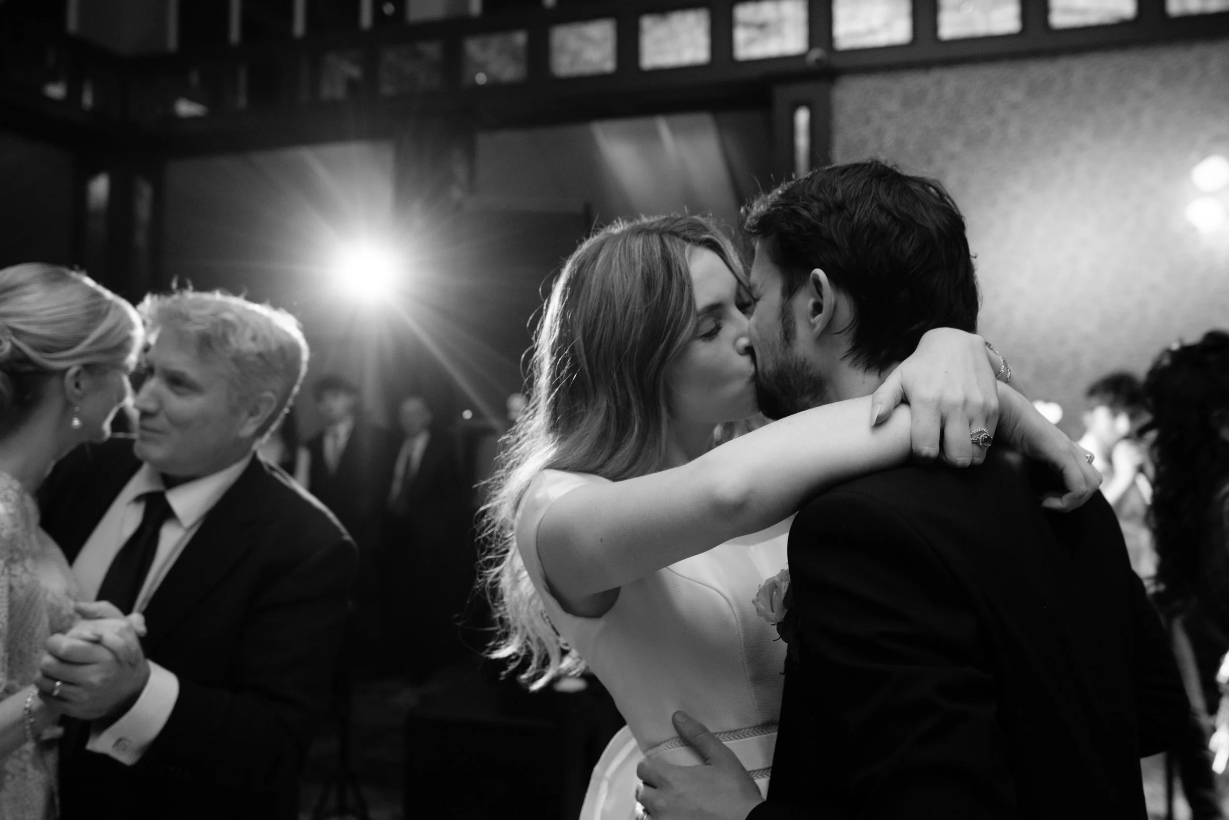 Black and white photo of a romantic moment at a wedding reception, with a couple kissing and dancing, while other couples dance in the background.